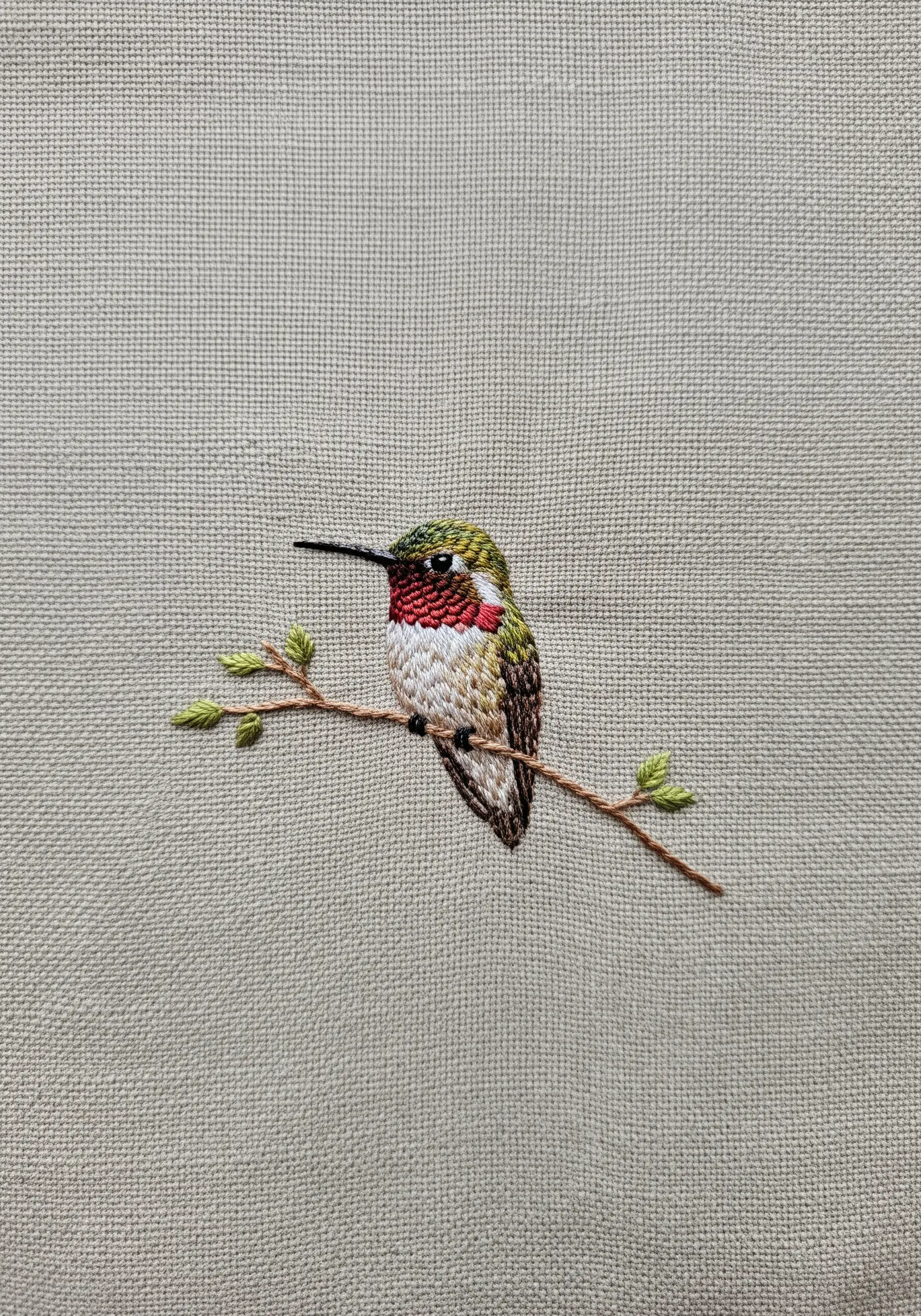 A single, small, detailed hummingbird perched on a branch on plain fabric.