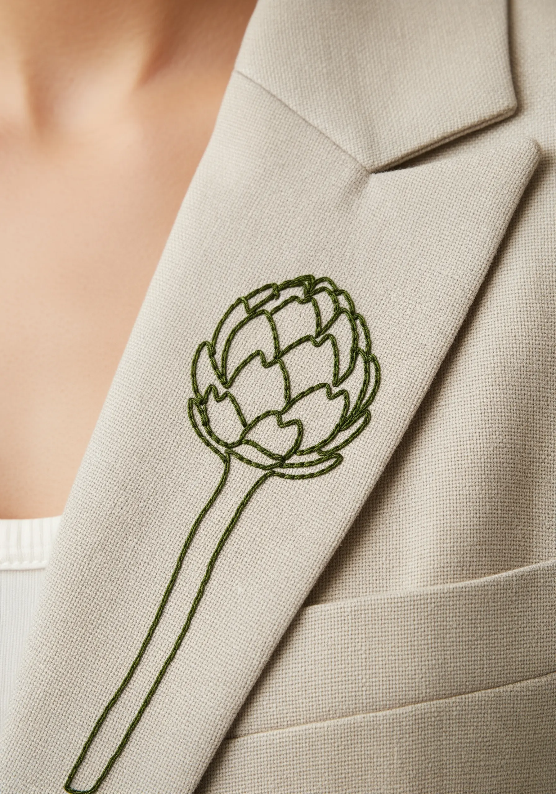 A single-line artichoke embroidered in dark green on the lapel of a beige blazer.