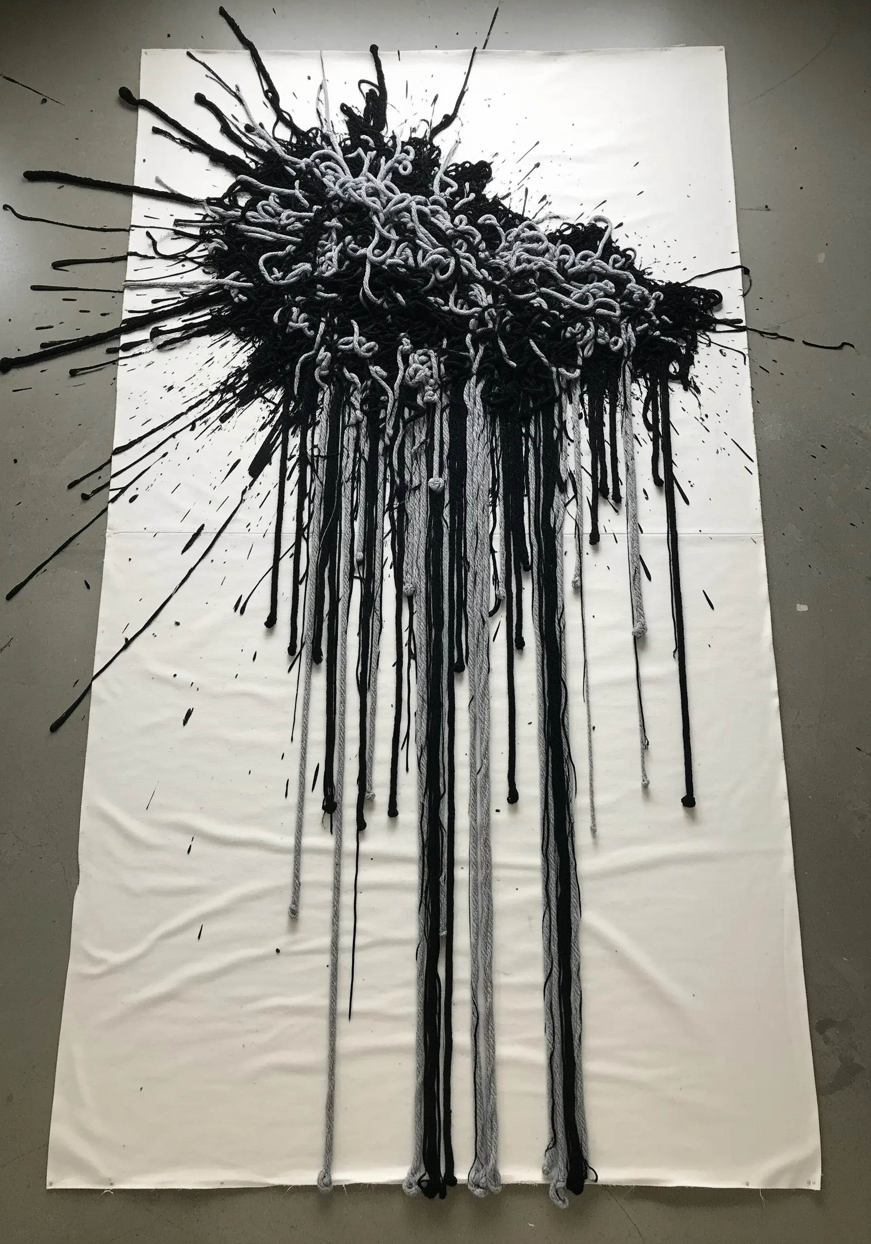 A large-scale fiber art piece with black and grey yarn dripping down a white canvas.