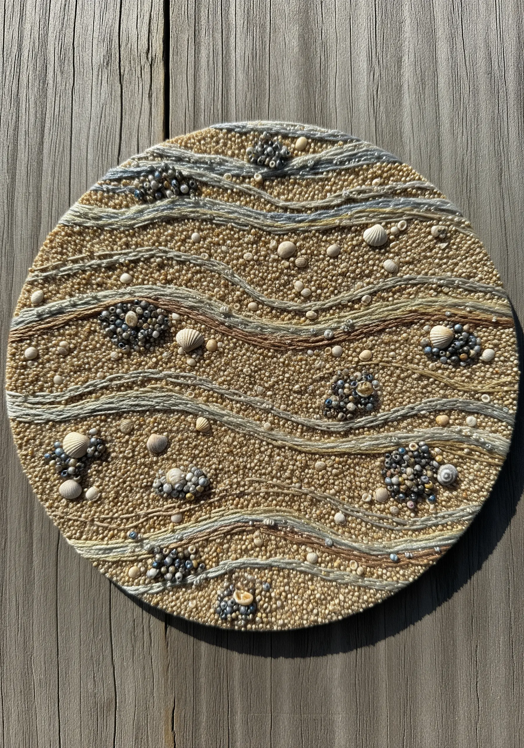 Circular mixed-media embroidery of a sandy beach with beads, shells, and French knots.
