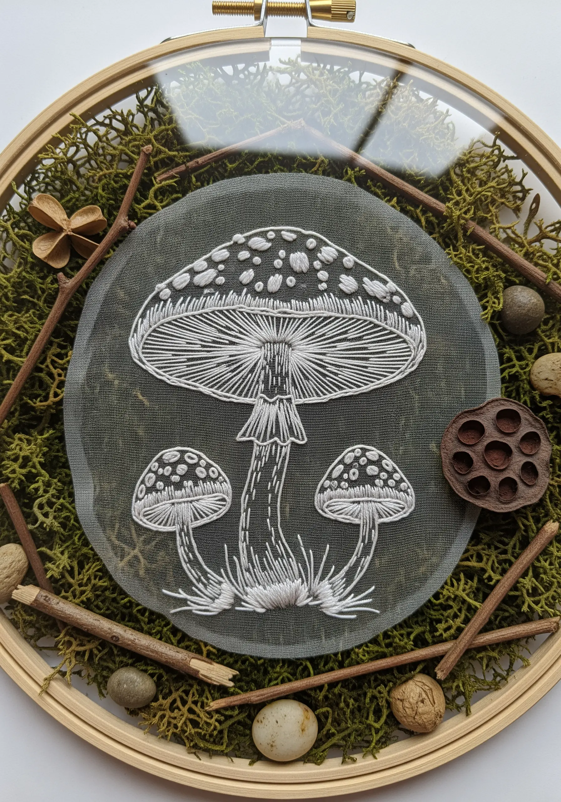 White line-art embroidery of mushrooms on sheer fabric, layered over moss and twigs.