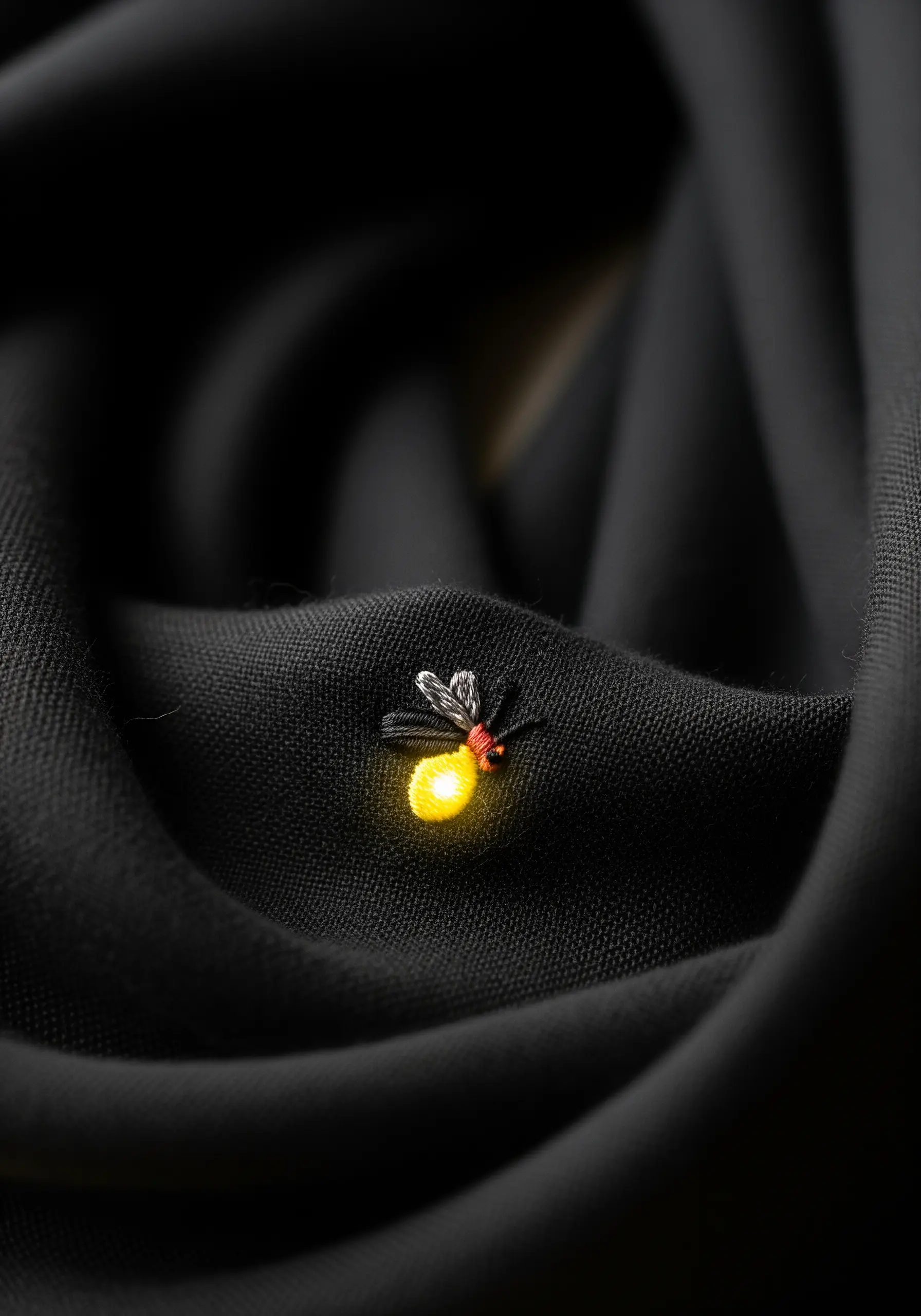 A tiny embroidered firefly with a glowing yellow abdomen on a black scarf.