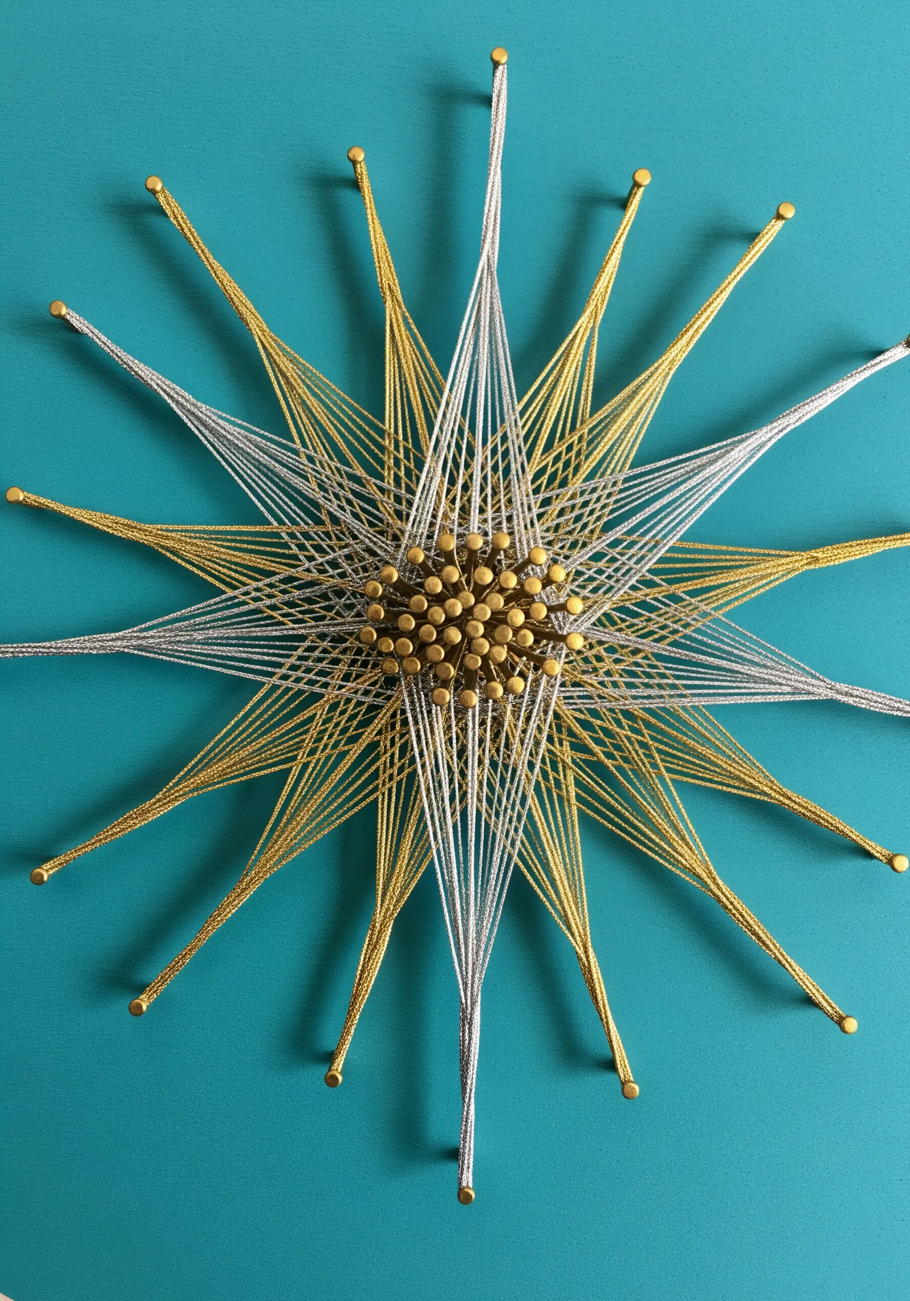 A starburst made of gold and silver string radiating from a central cluster of brass tacks