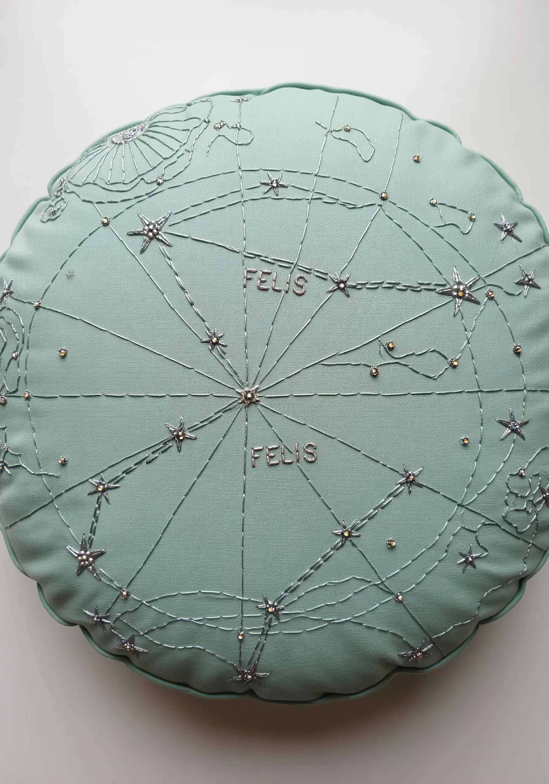 A celestial map embroidered with silver thread and beads on a round mint-green pillow.