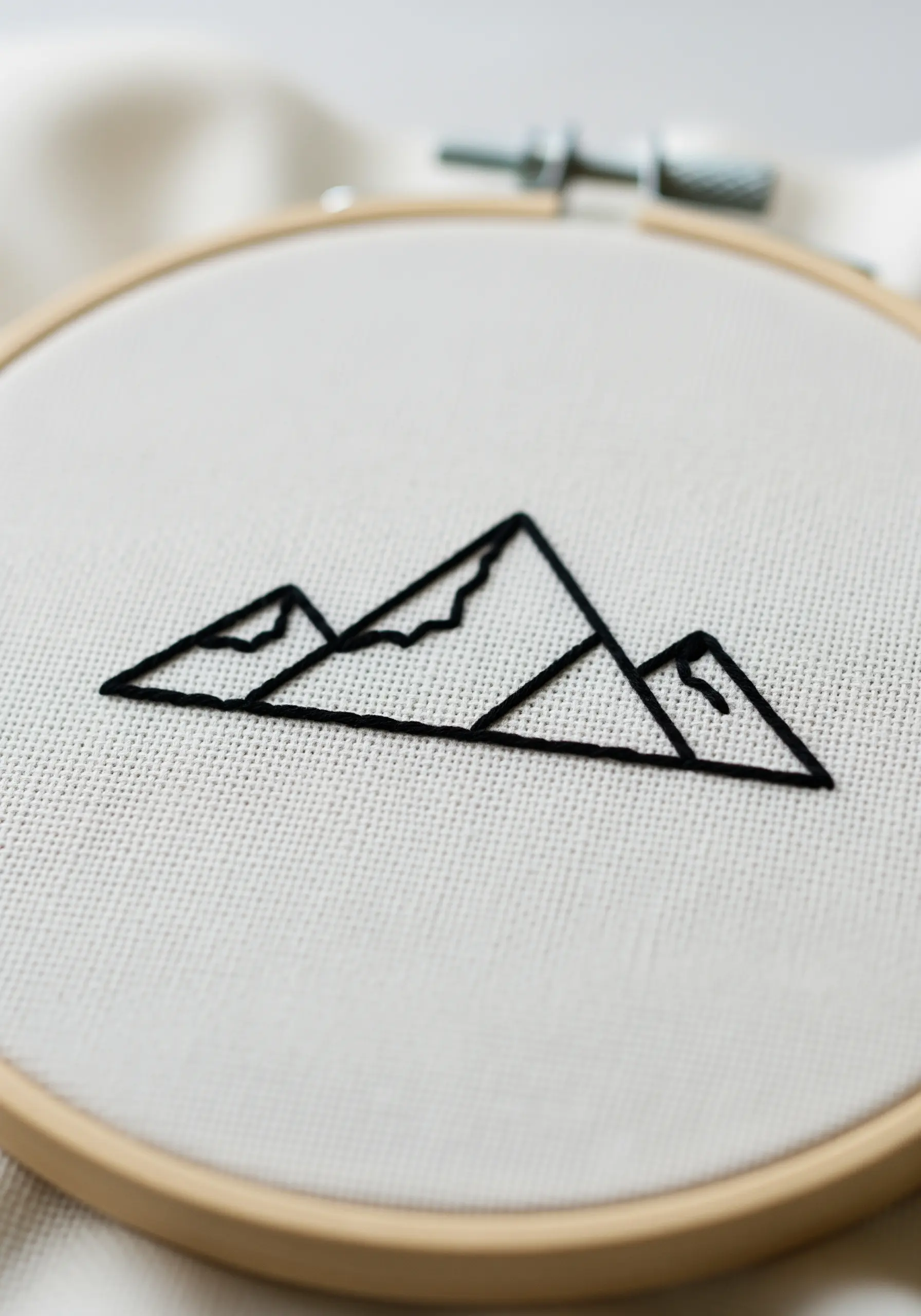Minimalist black line art of three mountain peaks embroidered on white canvas.