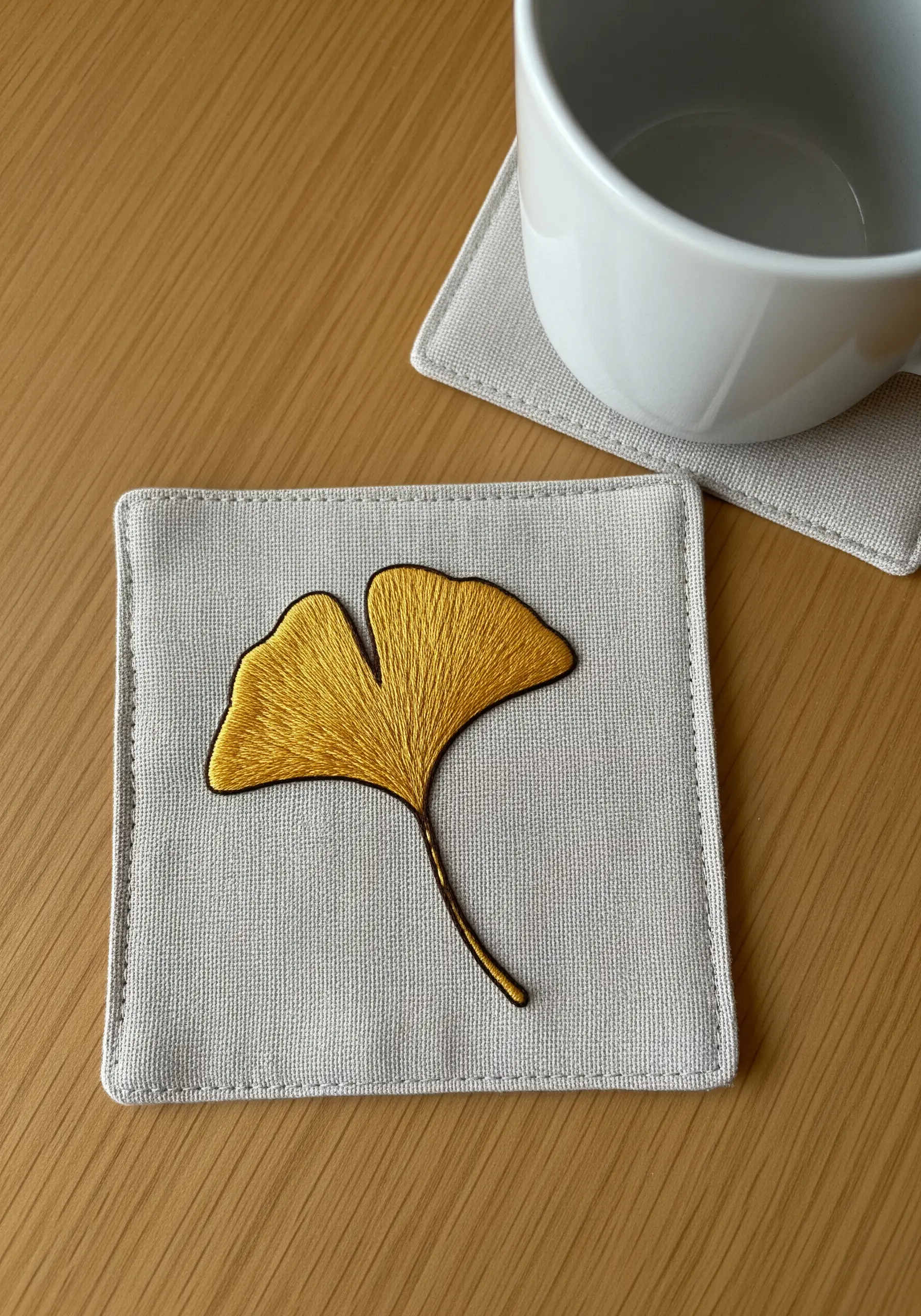 A golden ginkgo leaf with a smooth gradient fill stitch on a linen coaster.