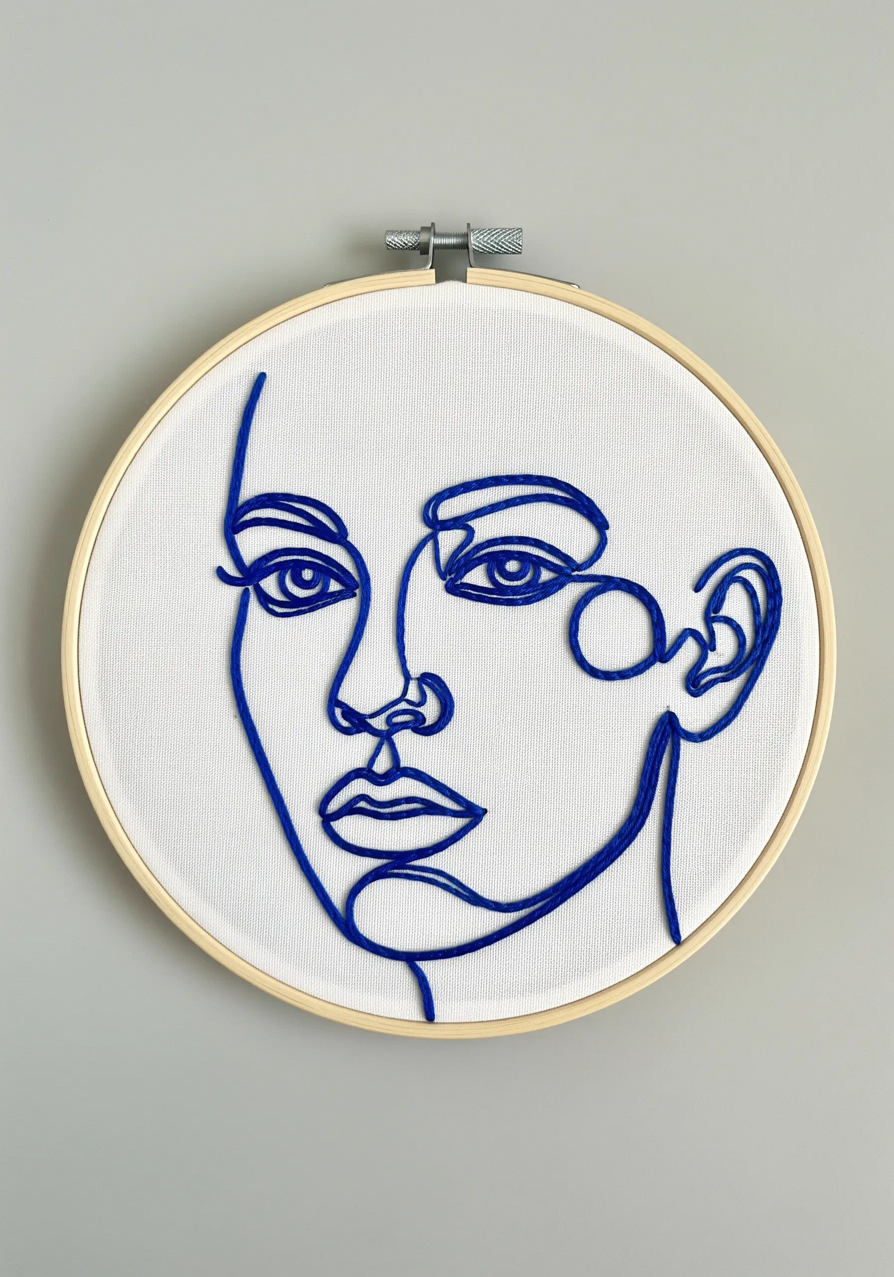 A continuous line art portrait embroidered in bold blue thread on white fabric.