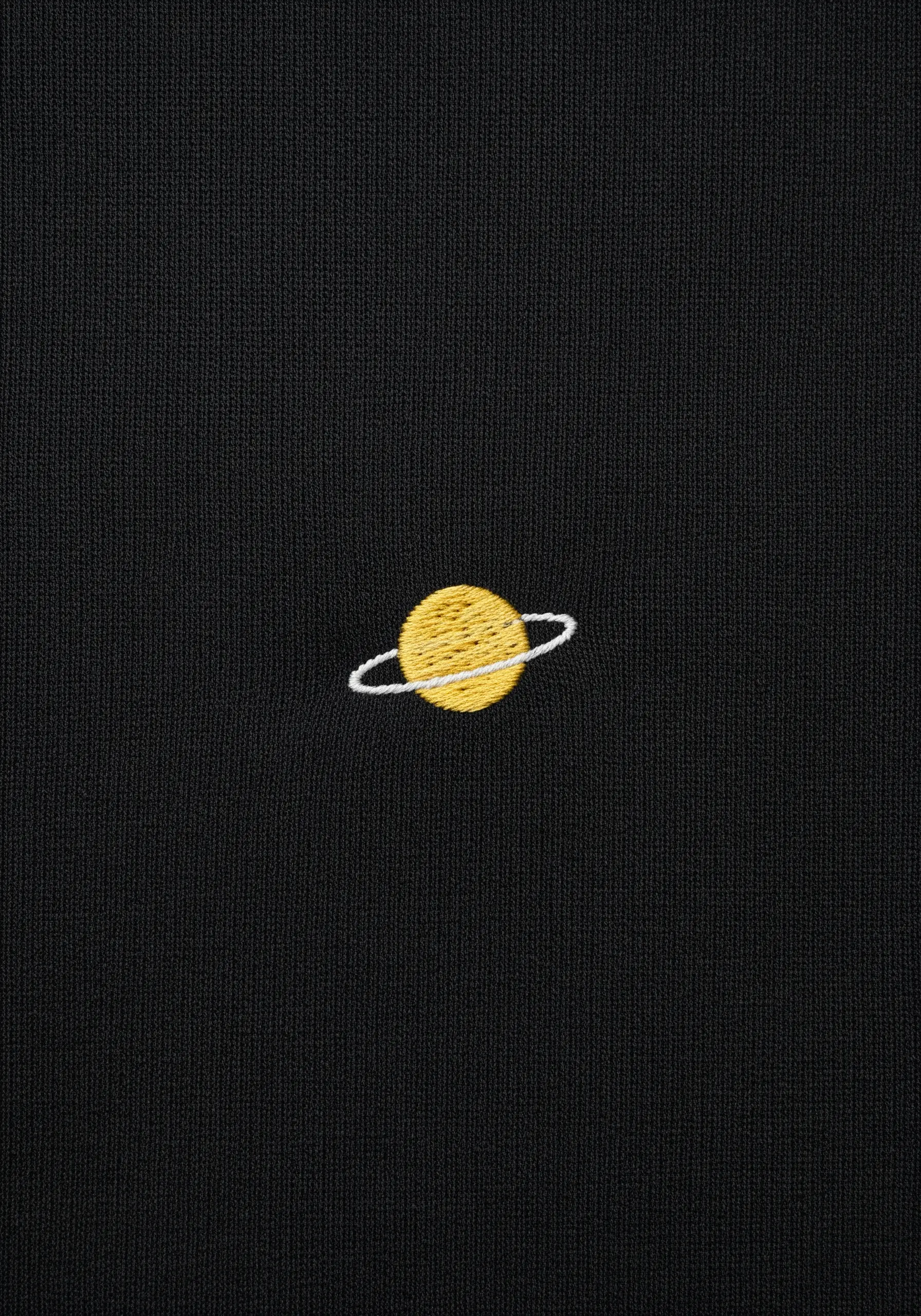 A tiny embroidered Saturn with a yellow satin stitch planet and white ring on black fabric.
