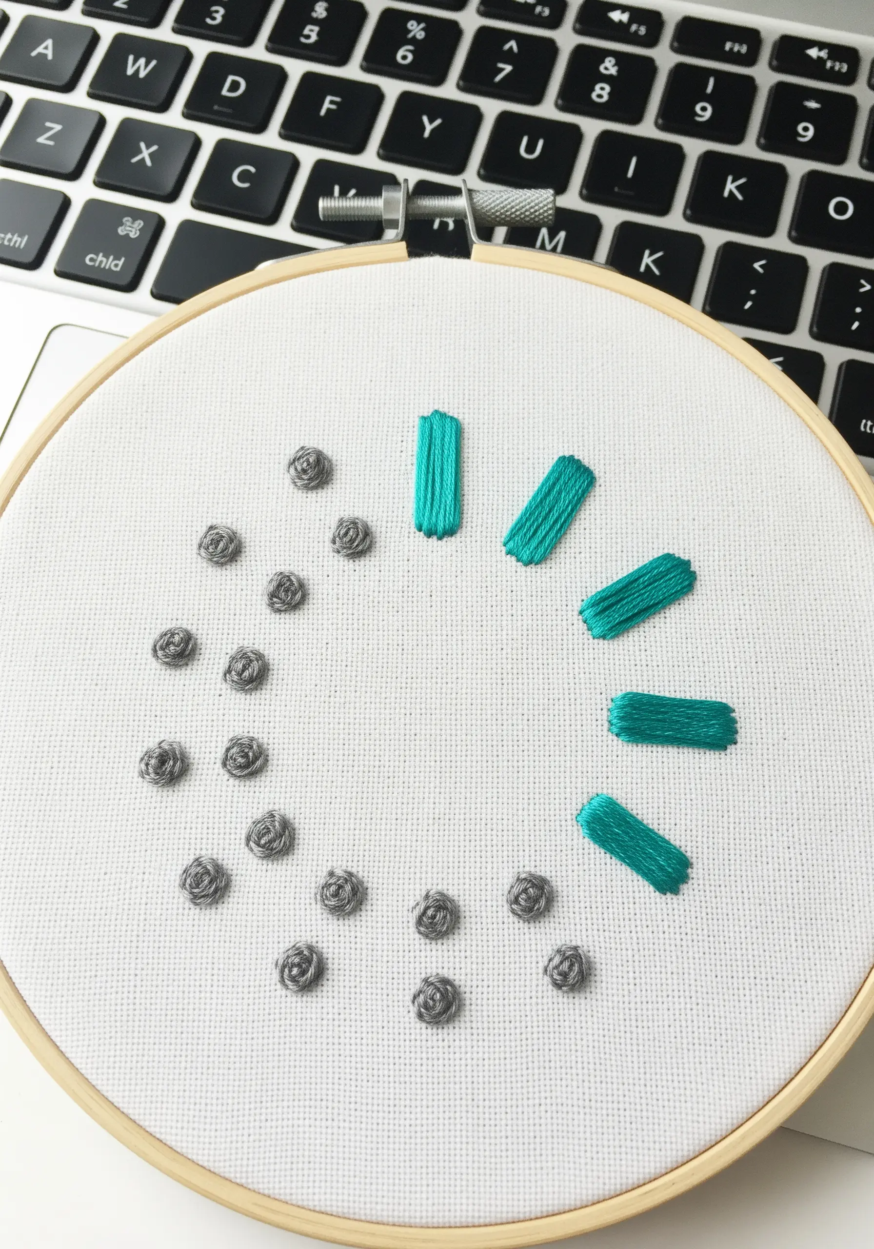 An abstract clock or loading icon with teal satin stitch bars and gray French knots.