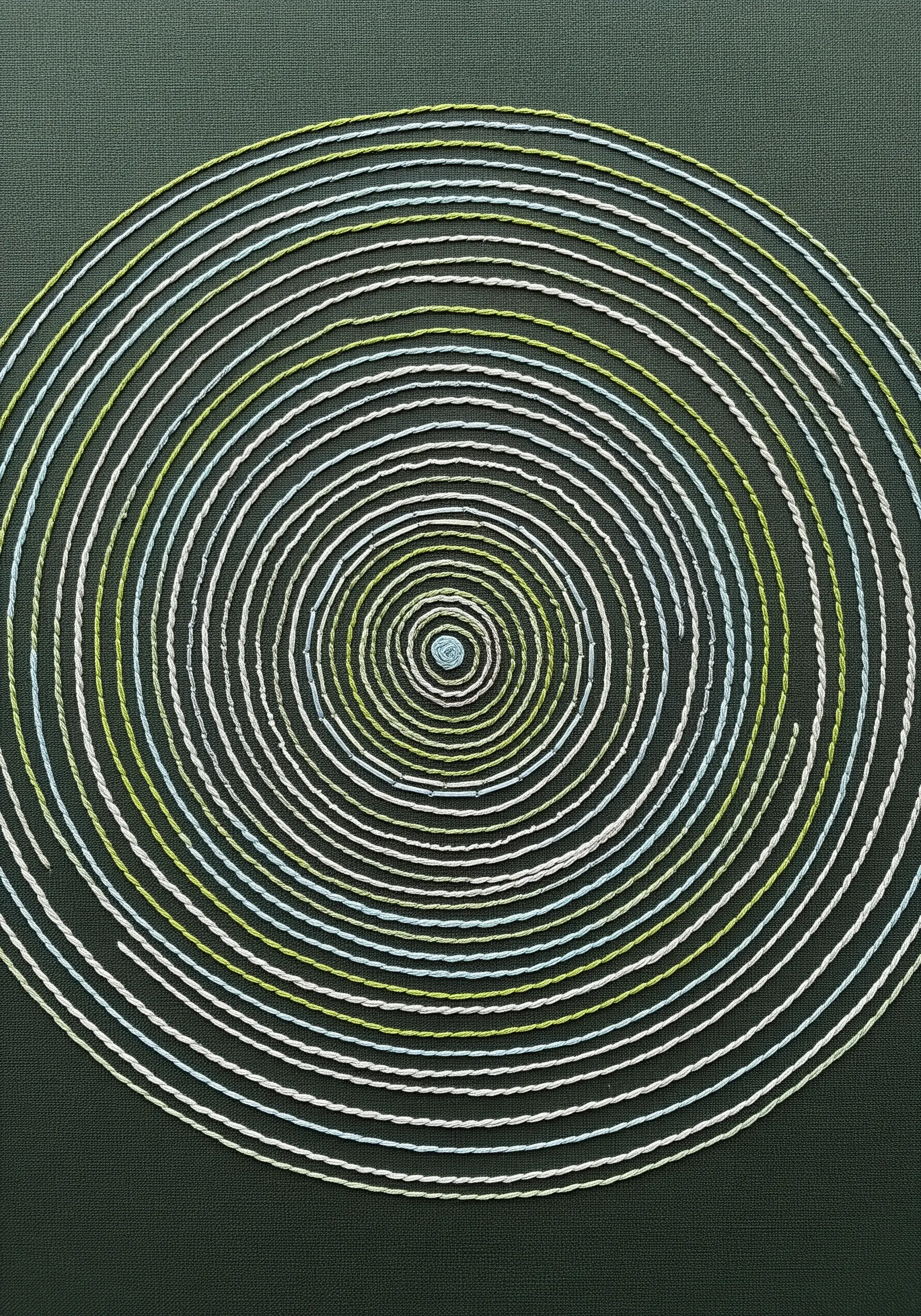 A simple embroidered spiral with white, green, and blue threads on dark fabric.