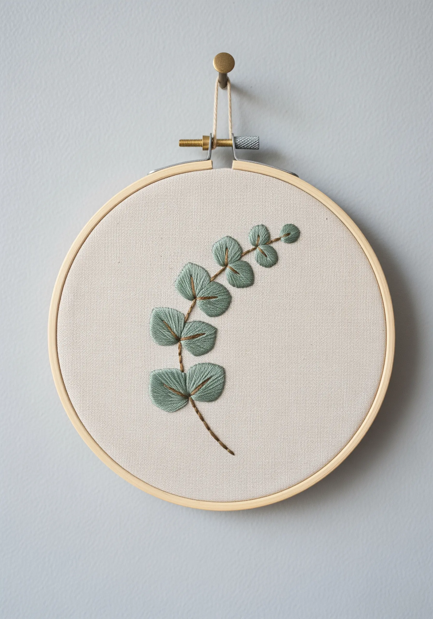 Embroidered eucalyptus branch with soft green leaves and brown outlines, hanging on a gray wall.