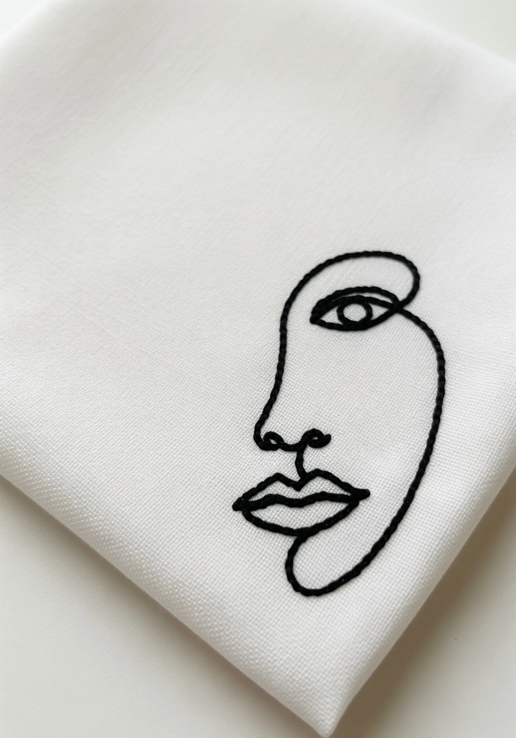 An abstract face embroidered with a single continuous black line on a white napkin.