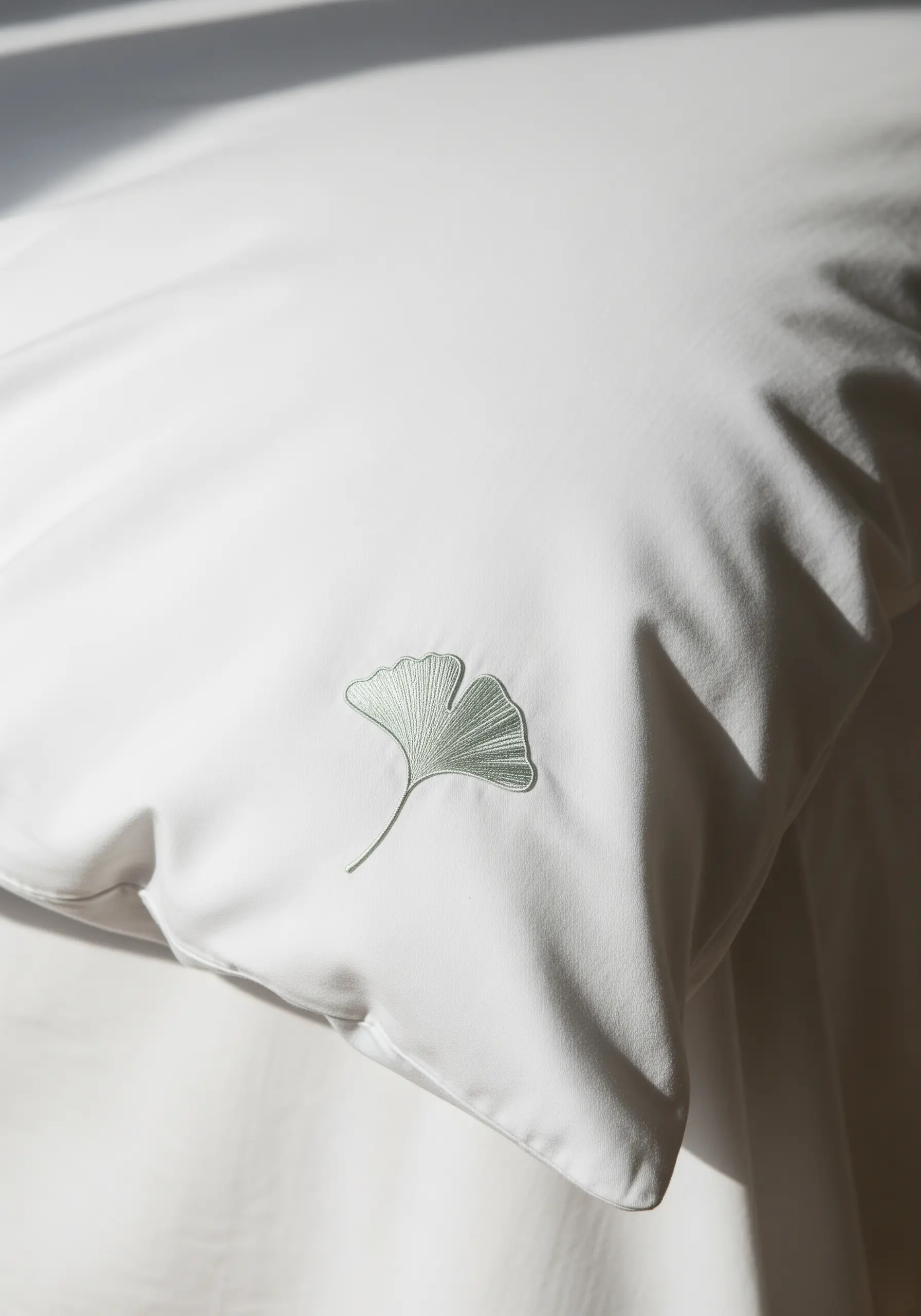 A single, beautifully executed satin stitch ginkgo leaf in pale green on a white pillowcase.