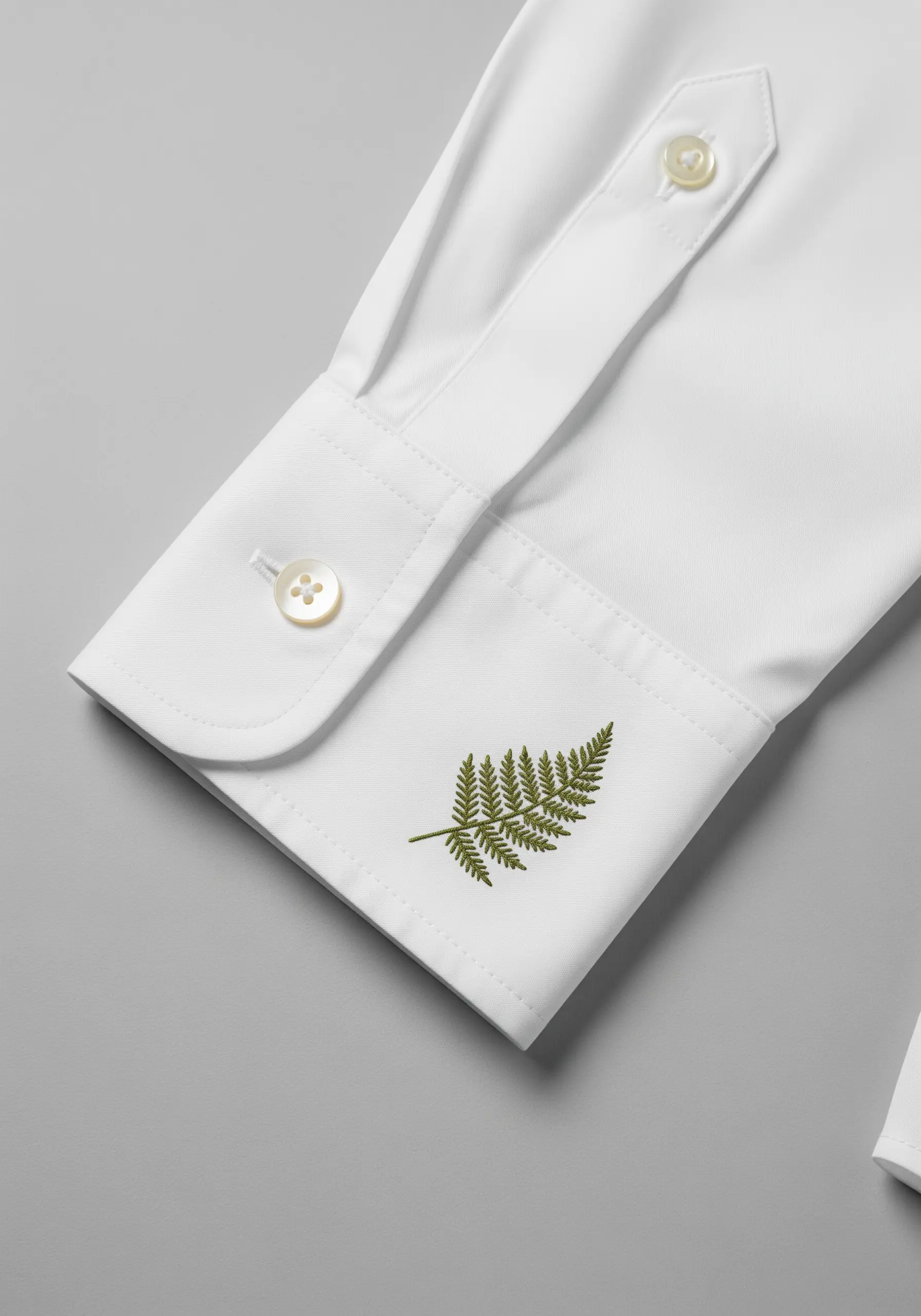 A detailed green fern leaf embroidered on the cuff of a white dress shirt.