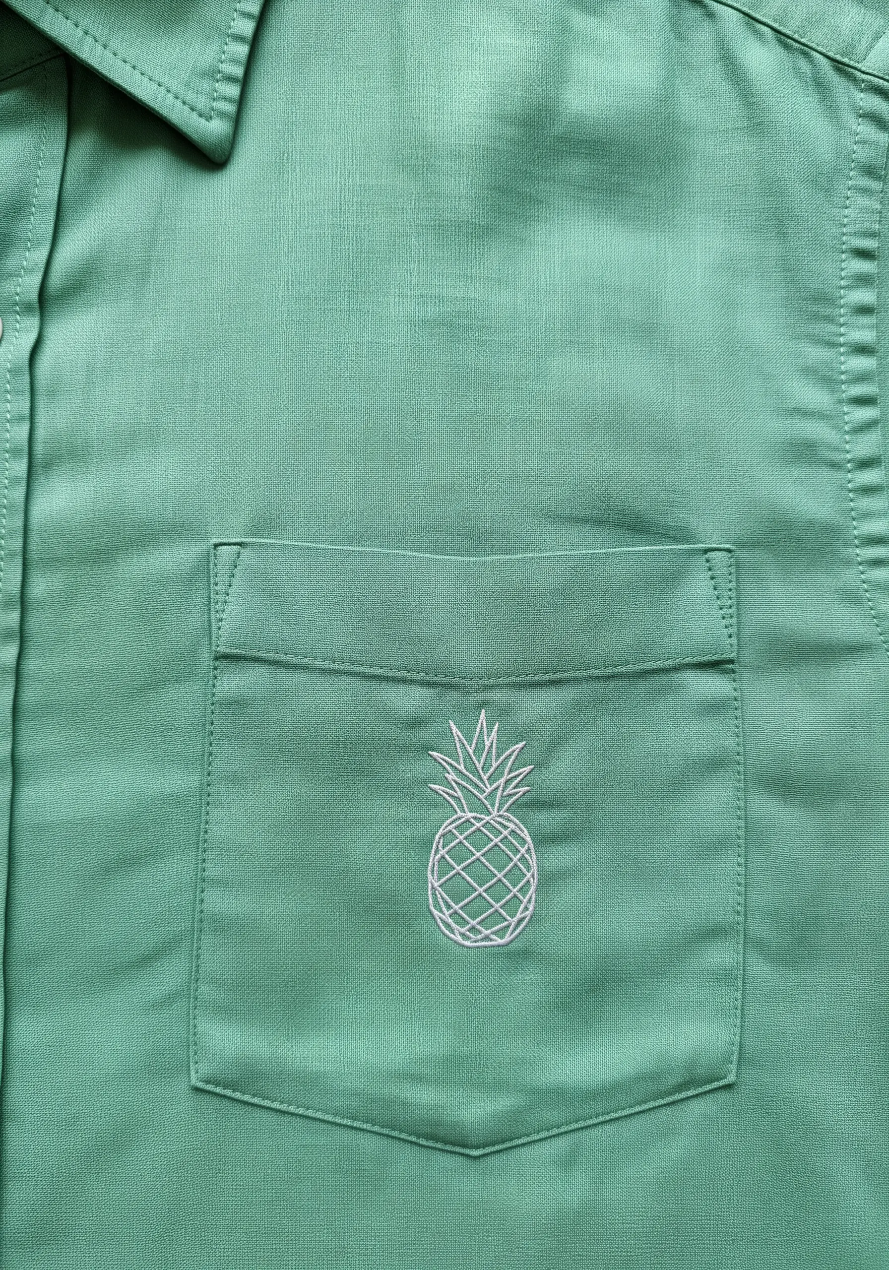 A minimalist white outline of a pineapple embroidered on a green shirt pocket.