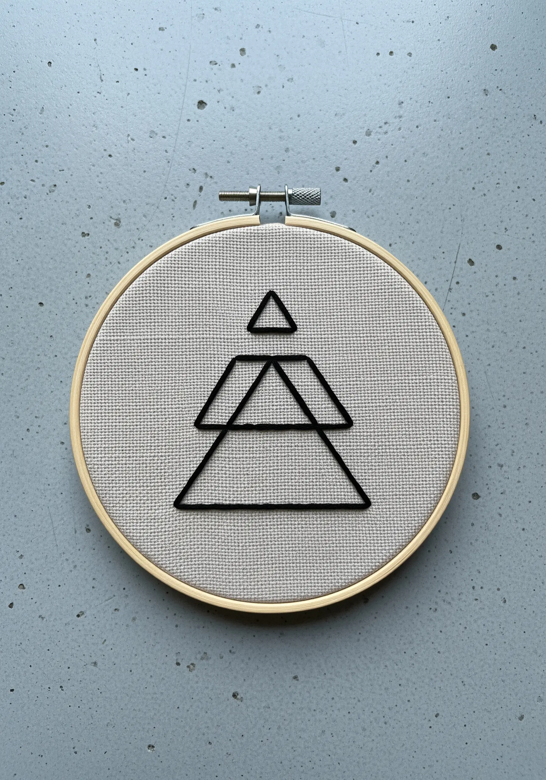 A crisp, black geometric symbol made of stacked triangles stitched on neutral Aida cloth.