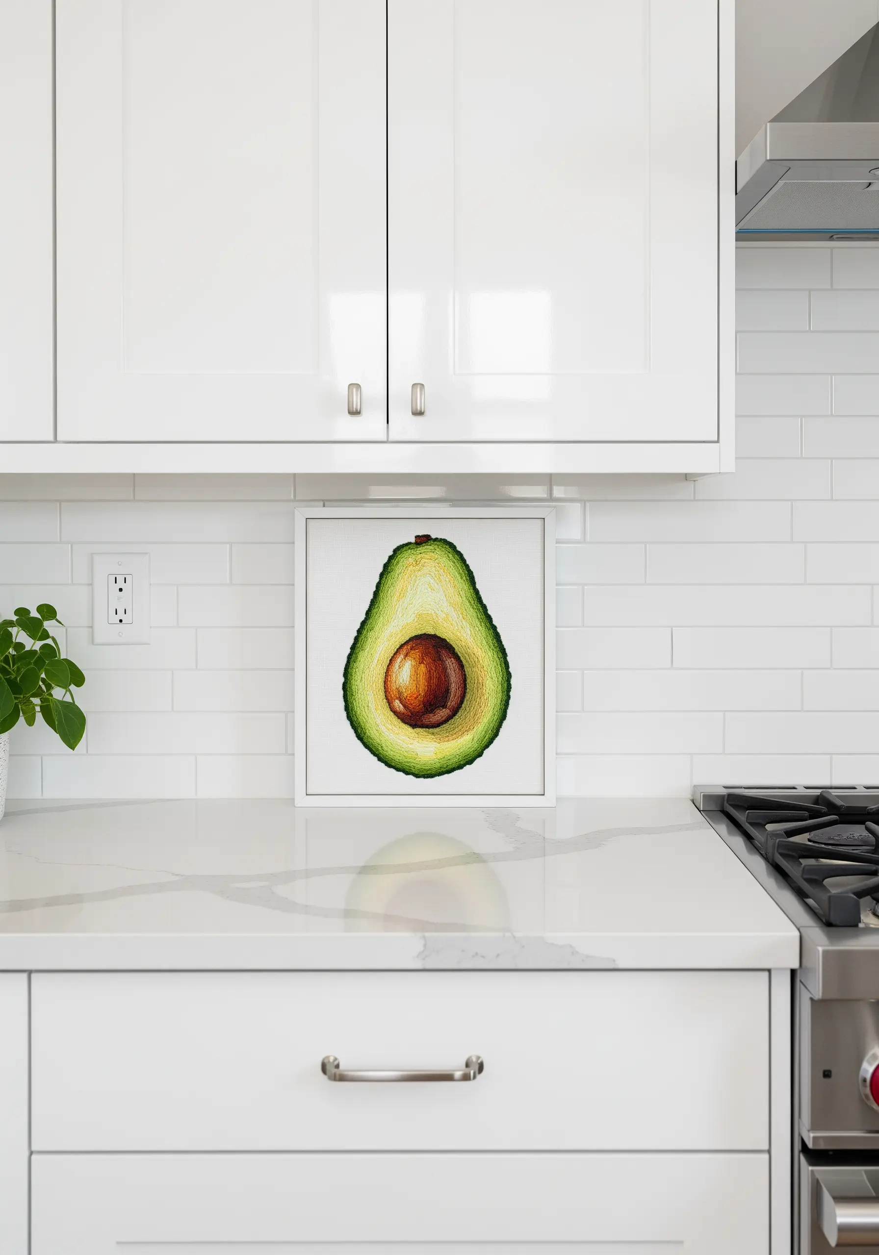 A framed, realistically embroidered avocado half sitting on a white kitchen counter.