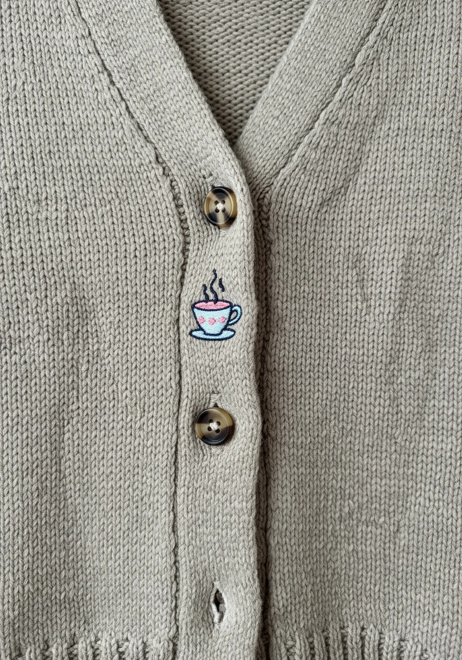 A tiny embroidered coffee cup taking the place of a button on a sage green knit cardigan.