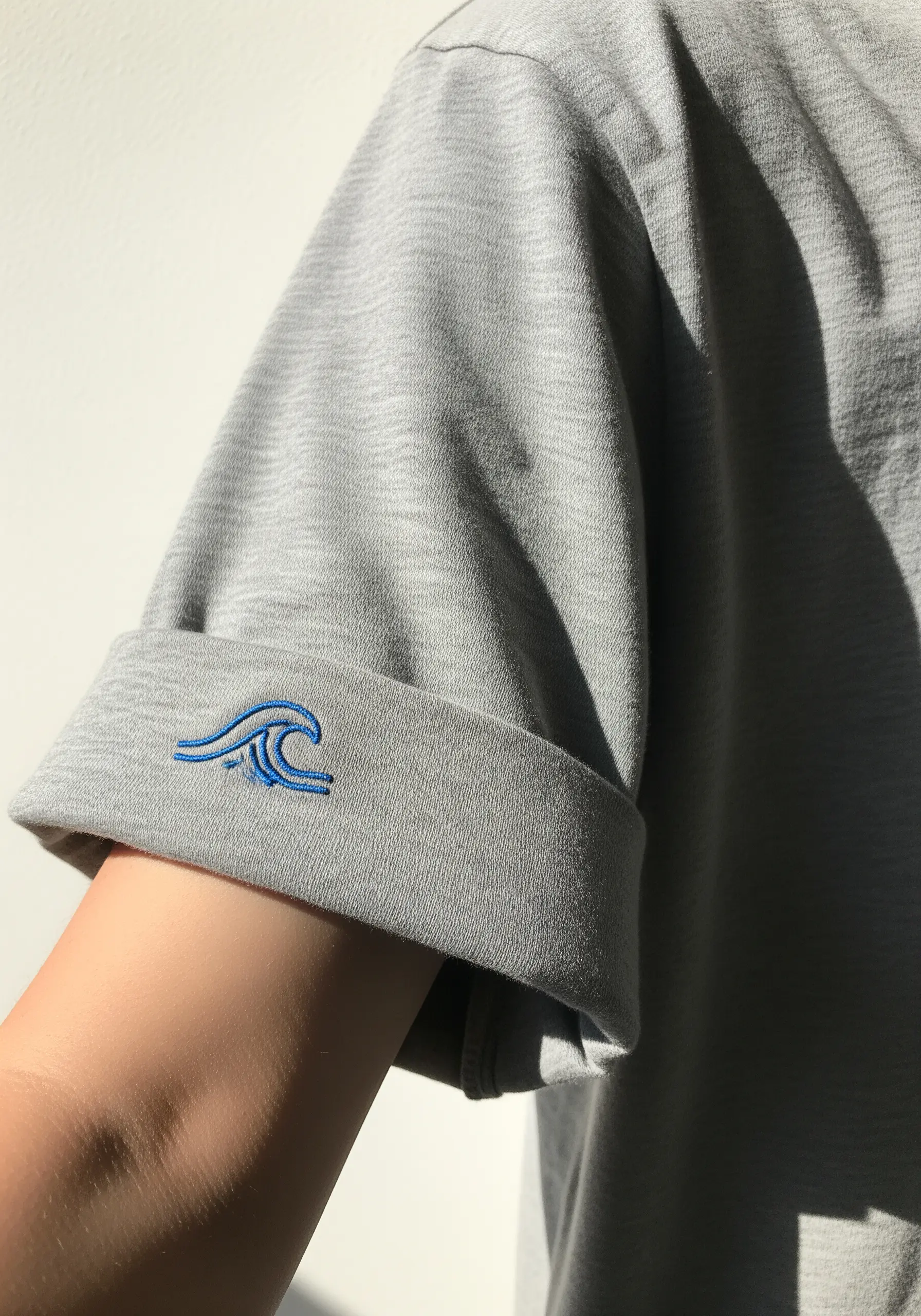 A simple, stylized blue wave embroidered on the rolled cuff of a grey t-shirt sleeve.