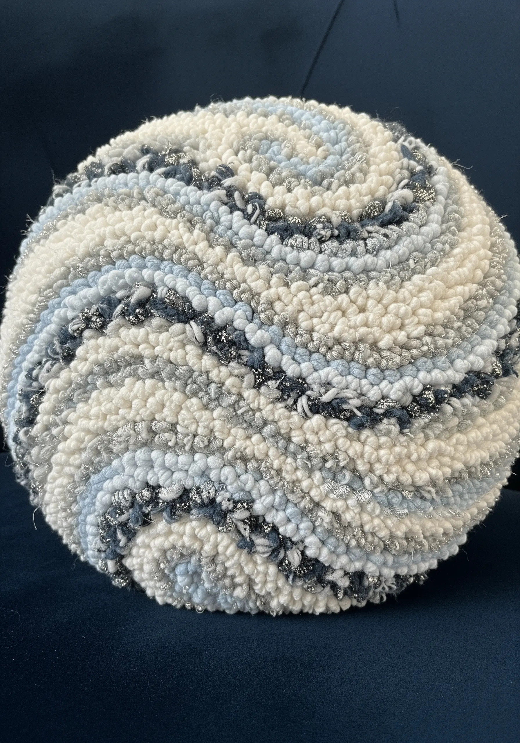 A spherical punch needle pillow with a swirl pattern in white, blue, and silver bouclé yarn.