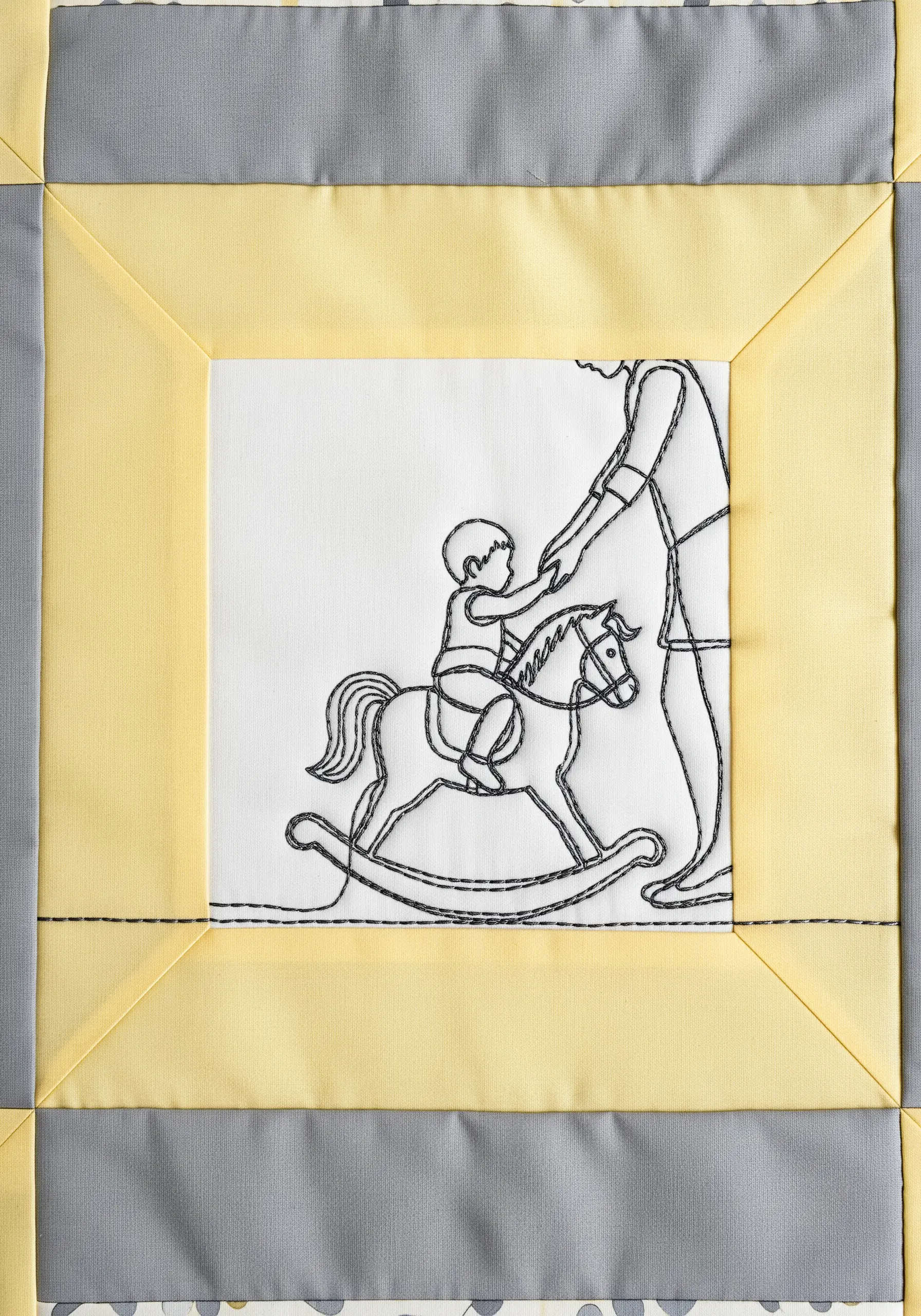 An embroidered line art motif of a child on a rocking horse, sewn into a yellow and grey quilt block.