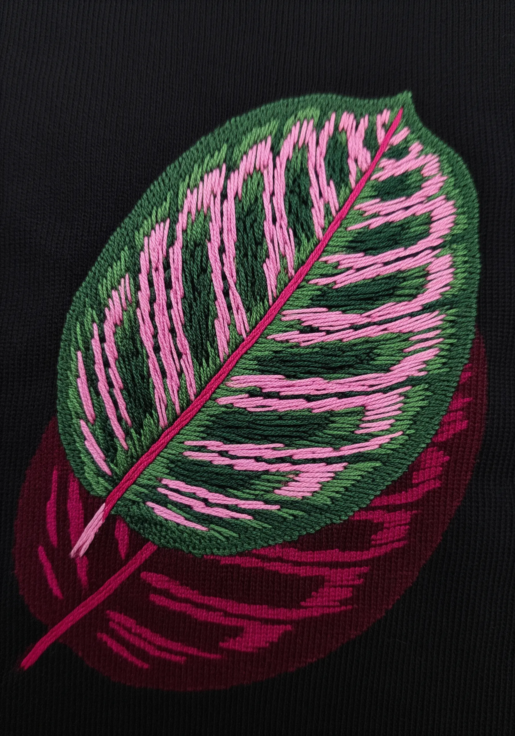 A green and pink calathea leaf embroidered on black fabric with a deep maroon shadow.