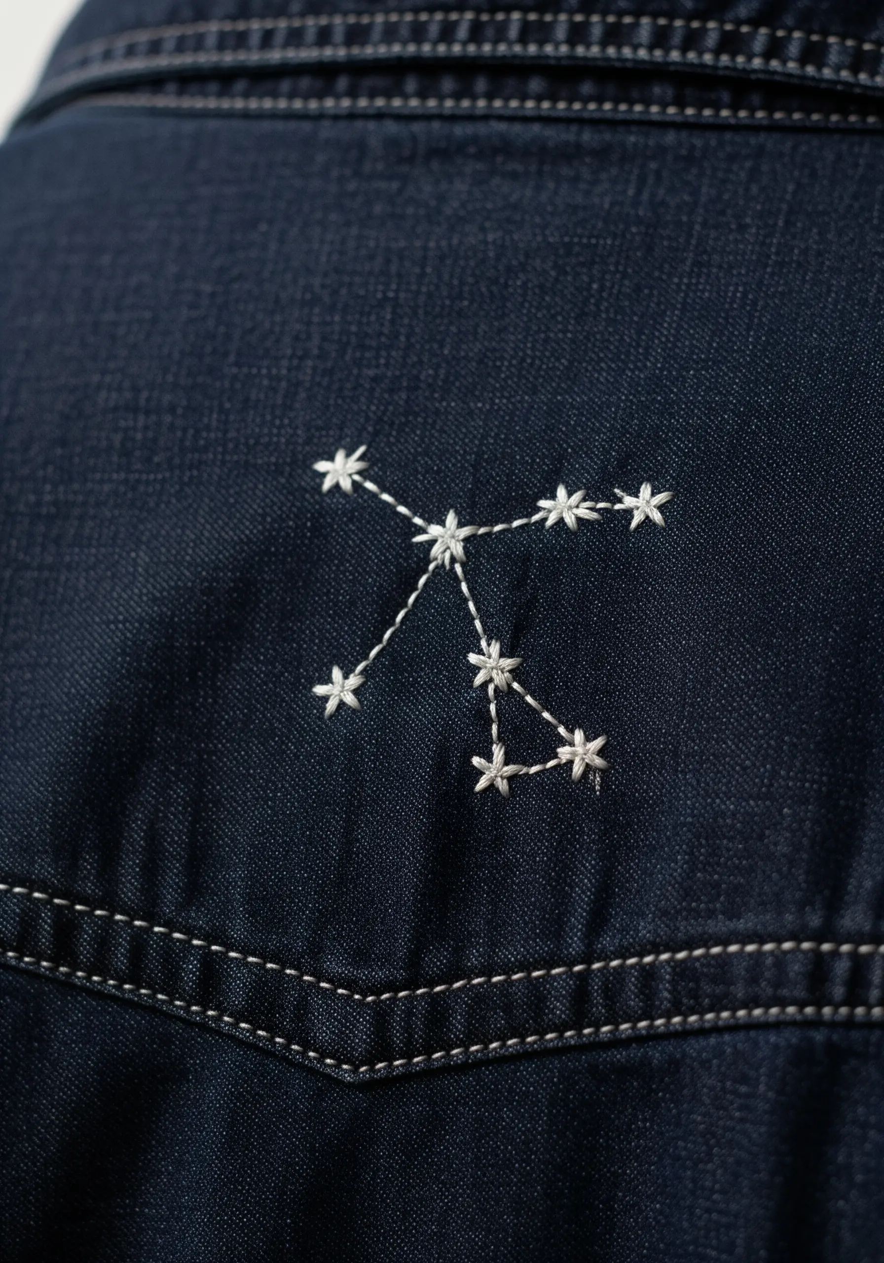 A small constellation stitched in white thread on the back pocket of dark denim jeans.