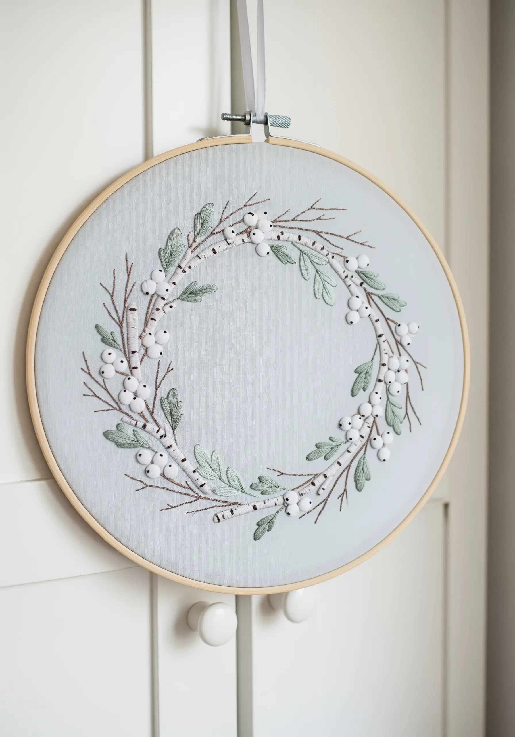 A winter wreath embroidery with white berries and birch branches on pale blue fabric.