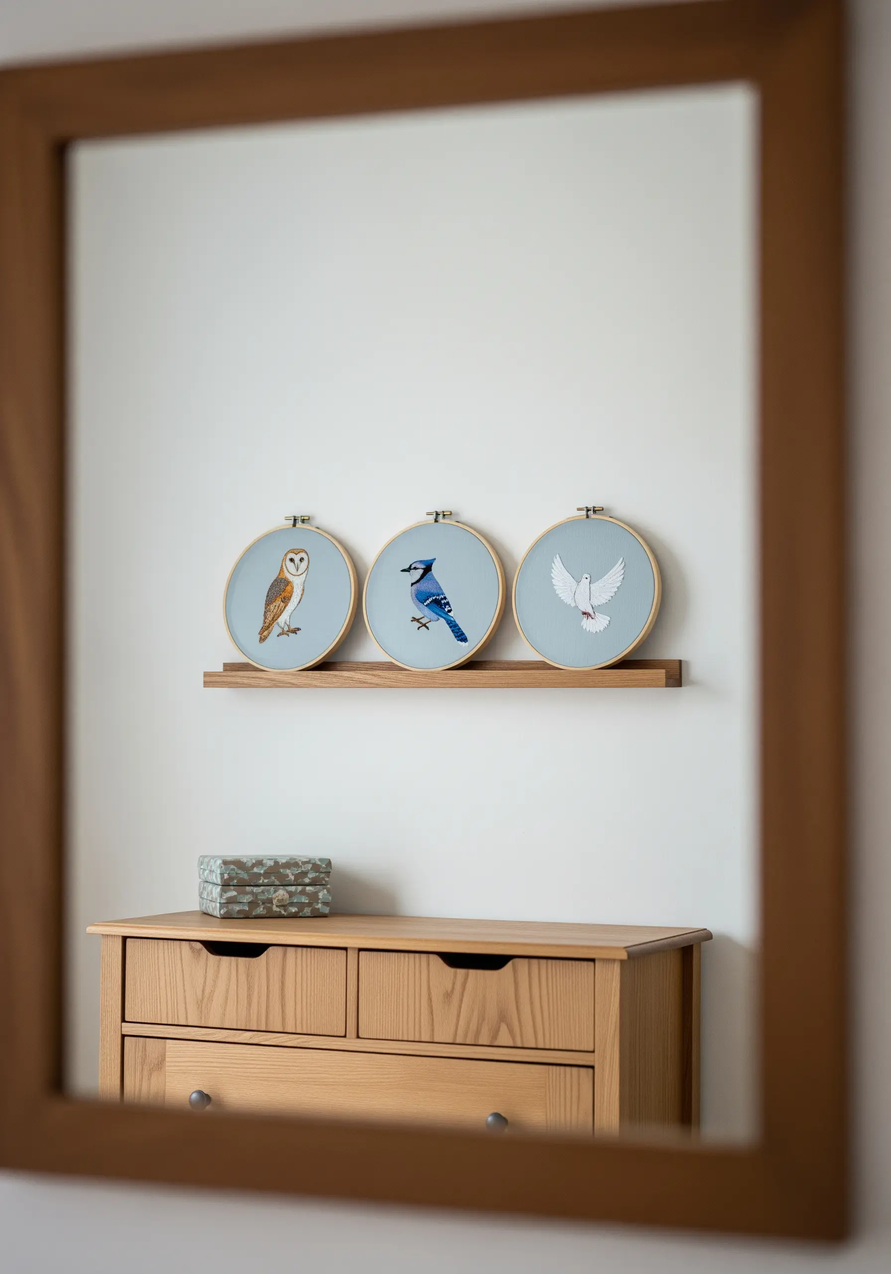 A trio of embroidery hoops on a shelf, each featuring a different realistically stitched bird.