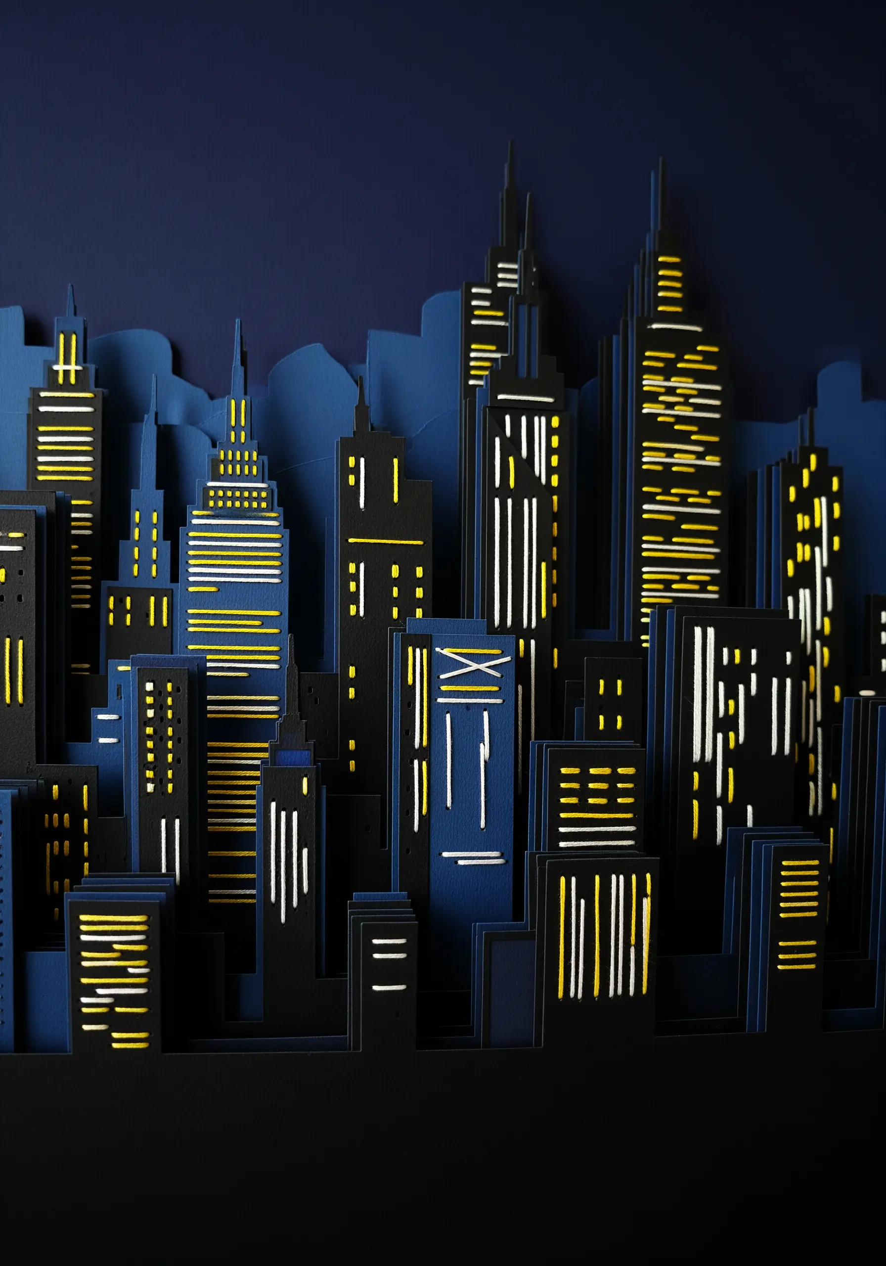 A layered paper-cut cityscape at night, with bright yellow thread stitched to represent lighted windows.