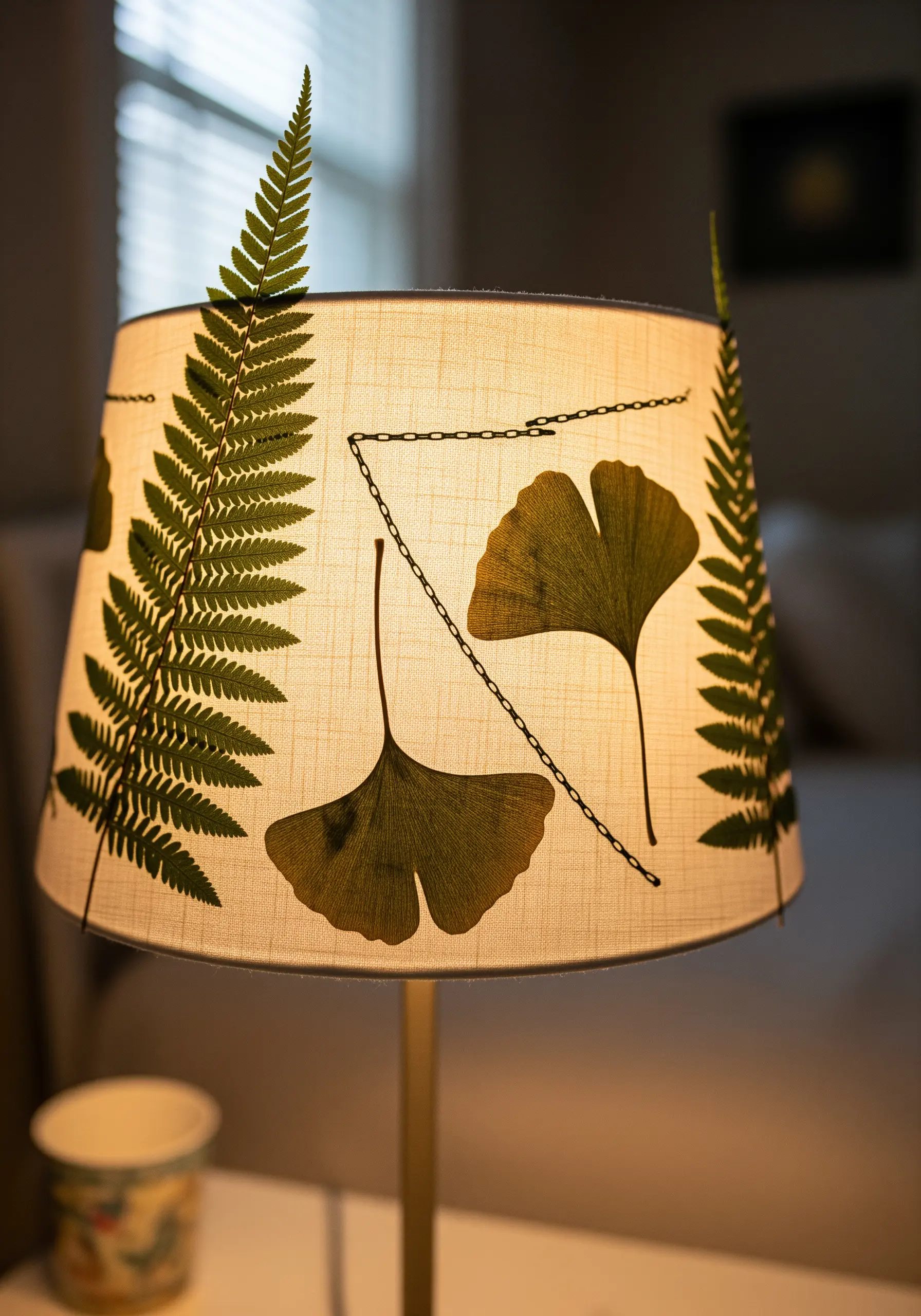A linen lampshade decorated with pressed fern and ginkgo leaves connected by a stitched chain.