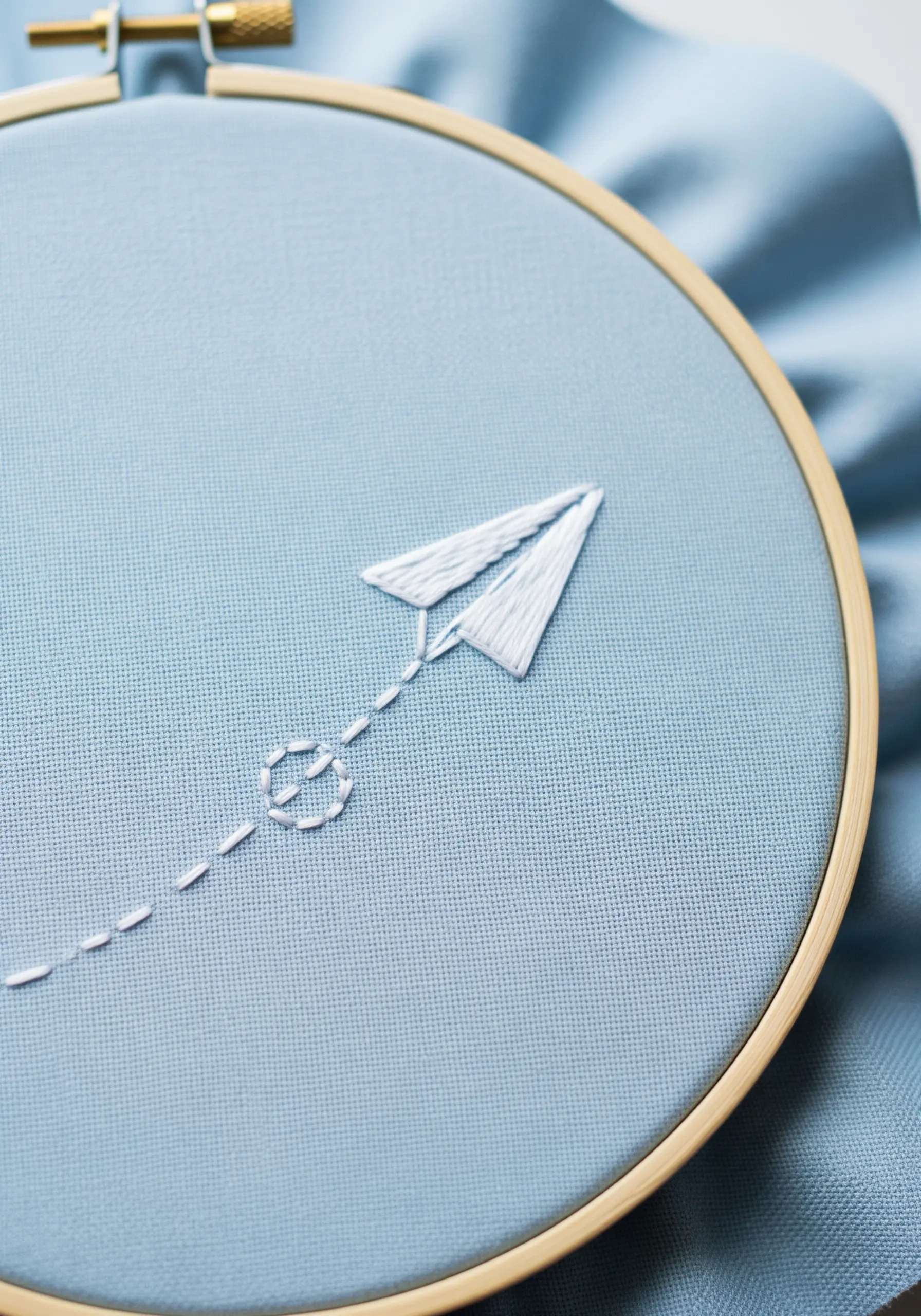 A white satin-stitched paper airplane with a dashed line of running stitches behind it.