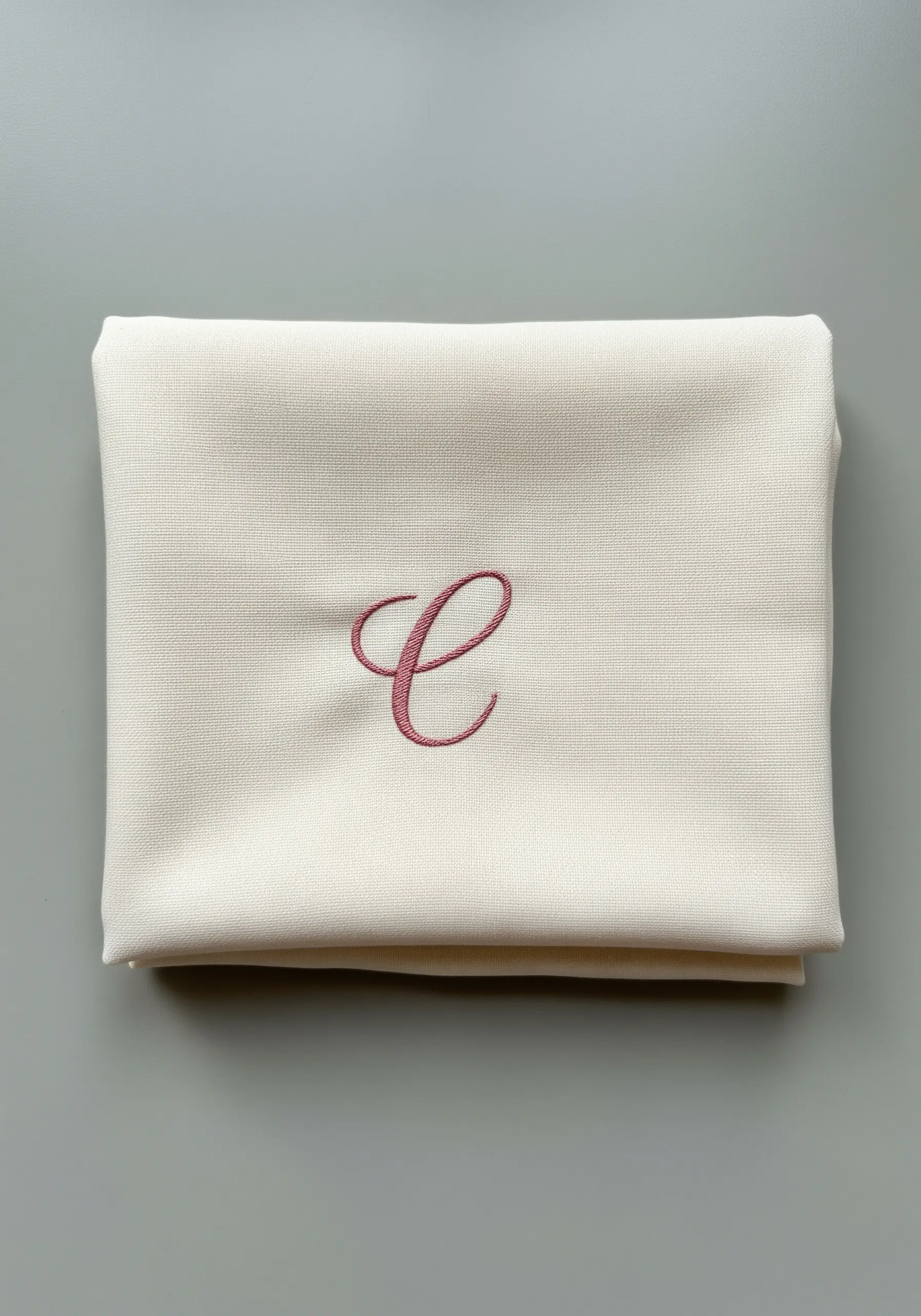 A single elegant cursive initial 'C' embroidered in dusty rose on a cream napkin.