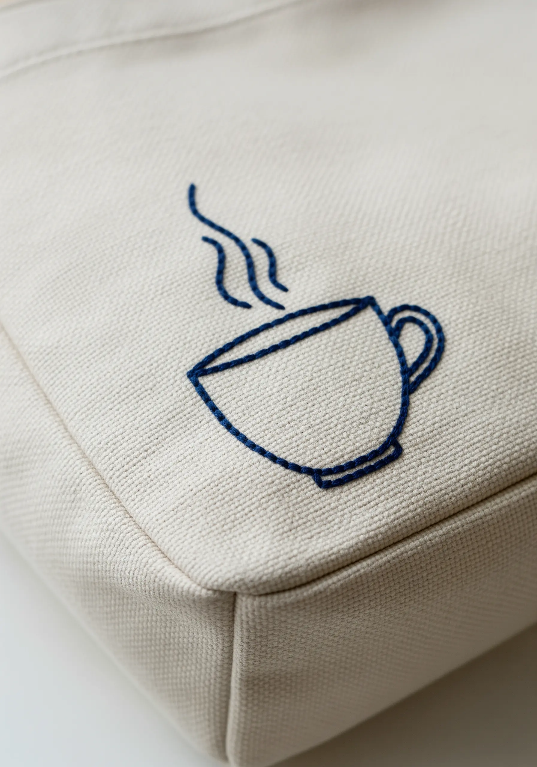 A simple line art embroidery of a steaming coffee cup on a canvas tote bag.