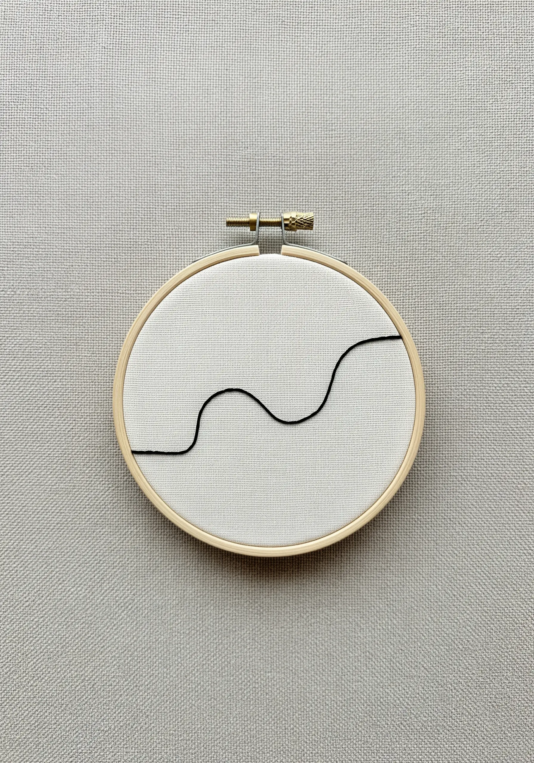 A single, minimalist black wavy line embroidered in a small hoop on light gray fabric.