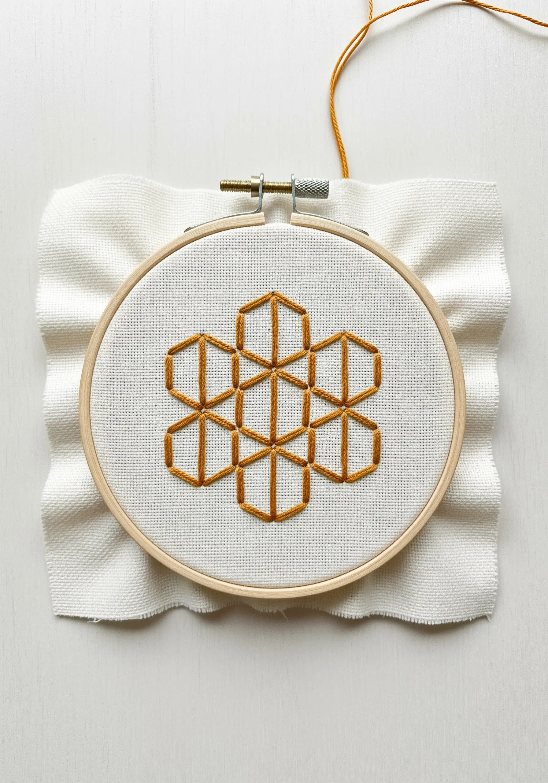 A geometric honeycomb pattern in ochre thread on white Aida cloth in a hoop.