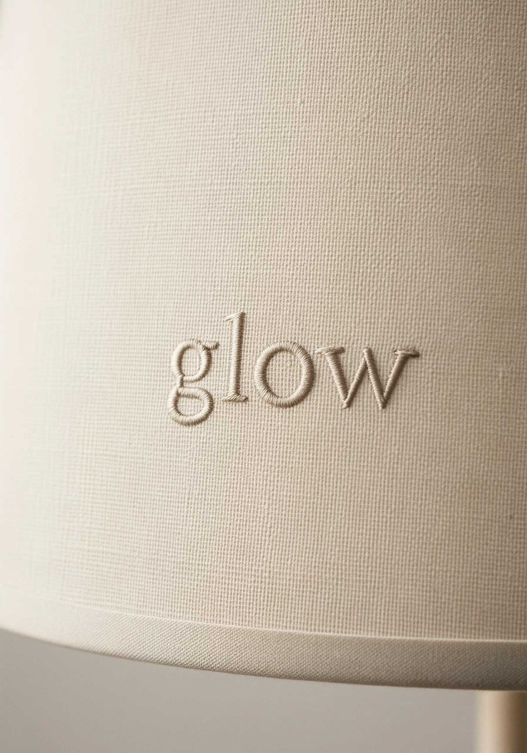 The word 'glow' embroidered in a light thread on a cream-colored fabric lampshade.