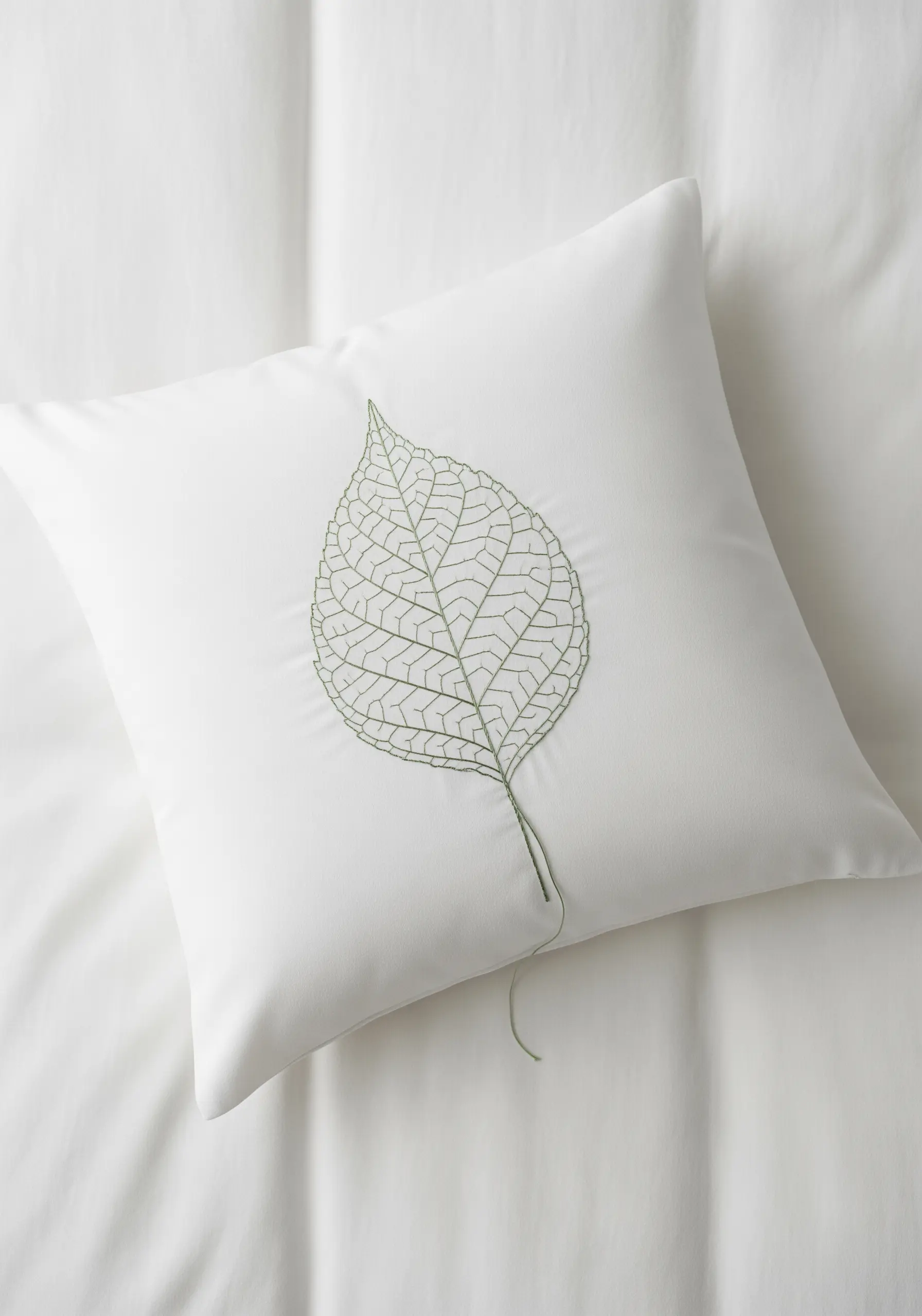 A detailed skeleton leaf embroidered in fine green thread on a crisp white pillow.