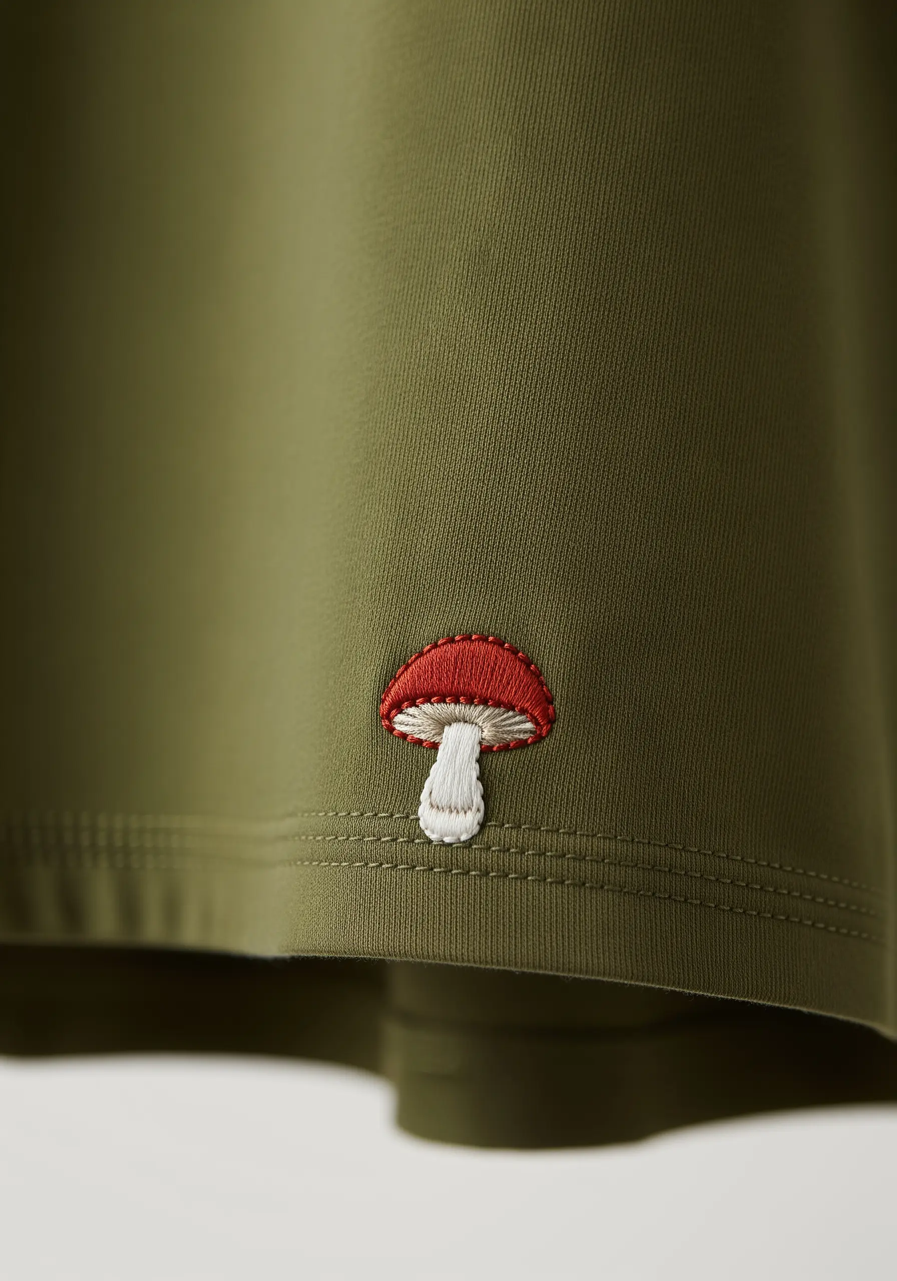 A tiny red and white mushroom embroidered just above the hem of an olive green shirt.