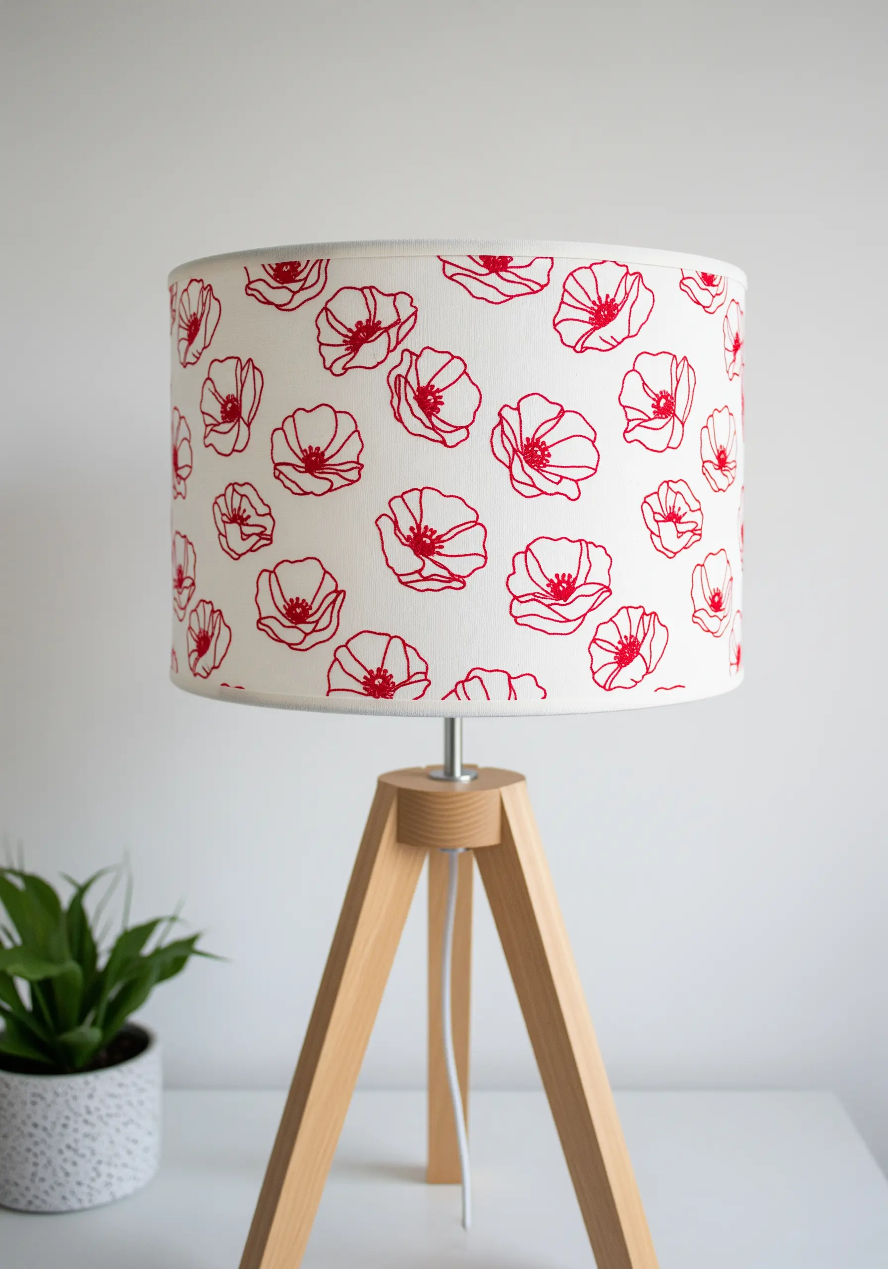 A modern tripod lamp with a white drum lampshade covered in red line-art poppy embroidery.