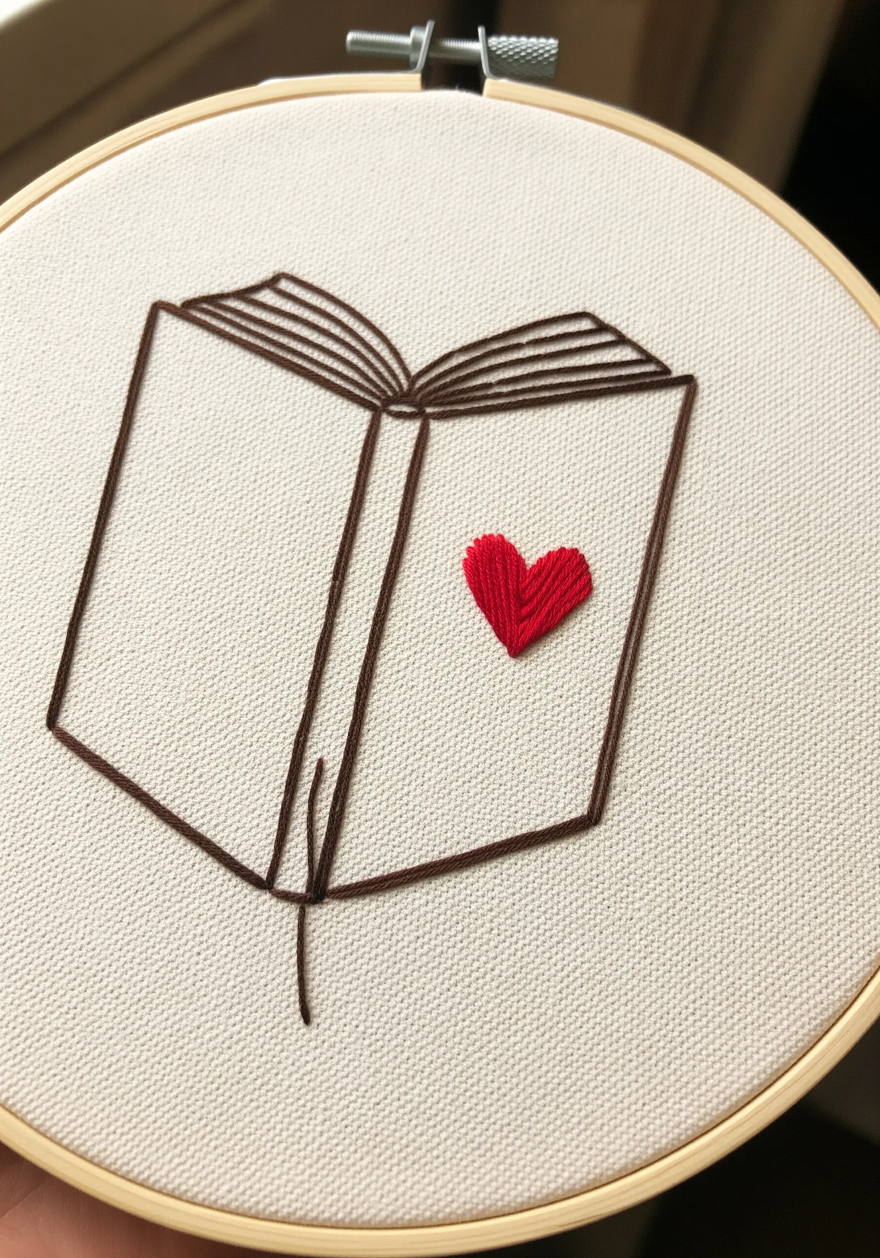 A line-art book embroidered in brown with a small, solid red satin-stitched heart.