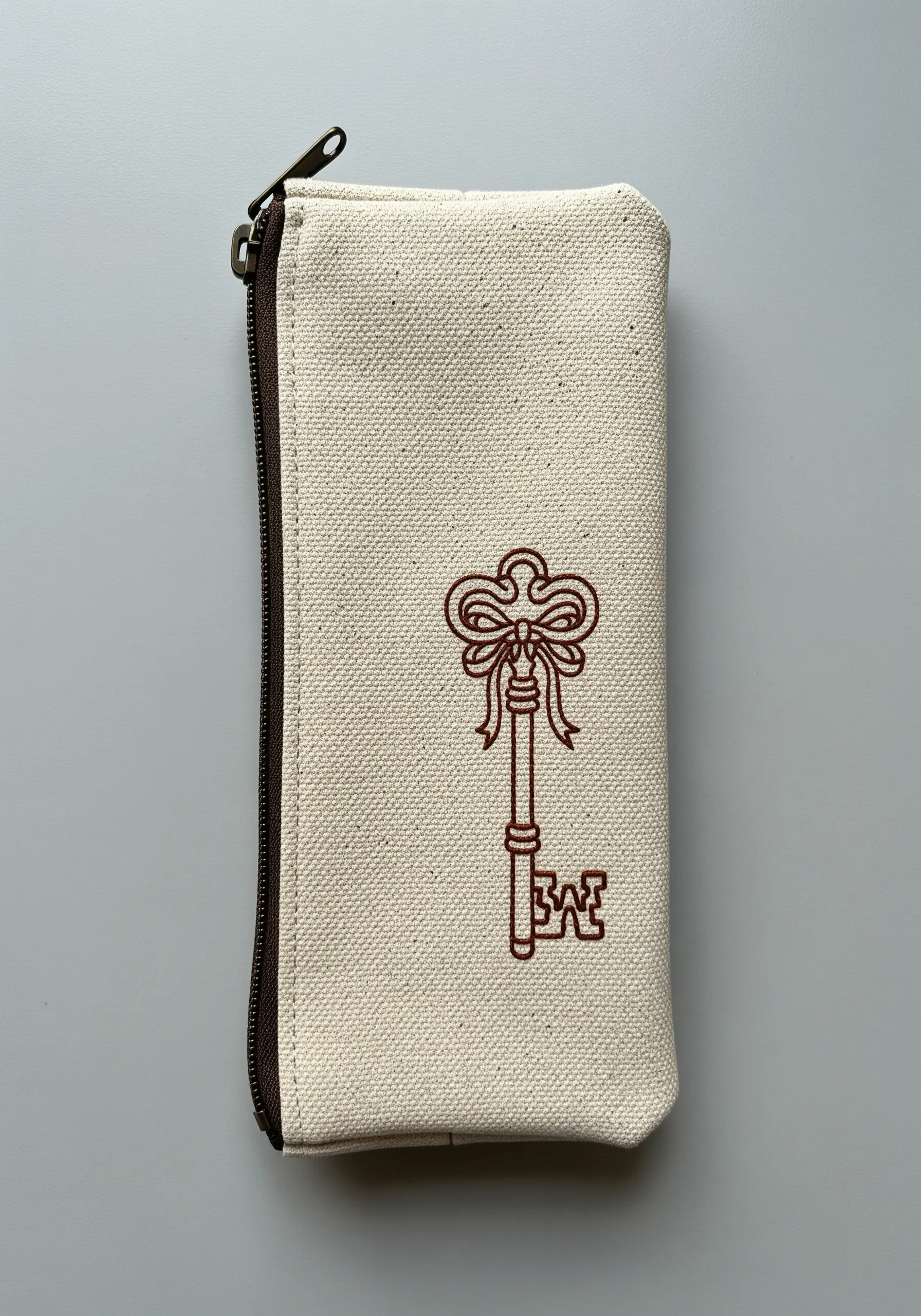 An ornate key with a bow embroidered in a thick brown outline on a canvas pouch.