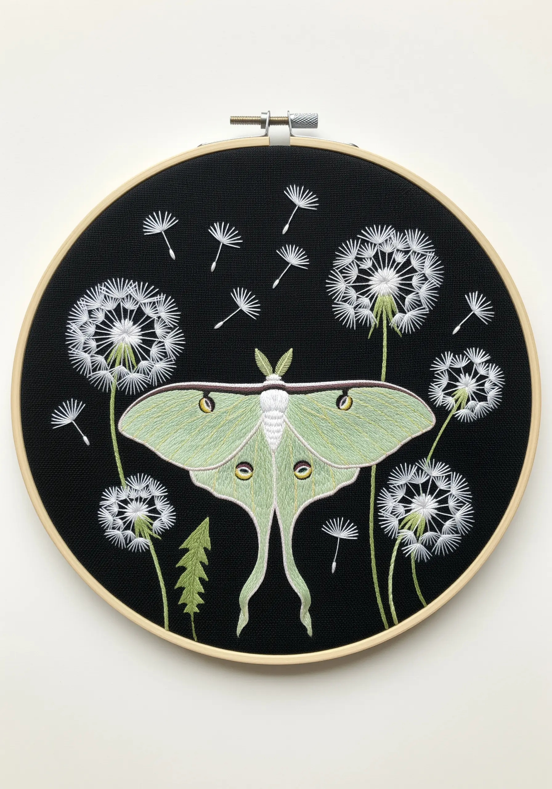 A pale green Luna Moth surrounded by floating dandelion seeds embroidered on black fabric.