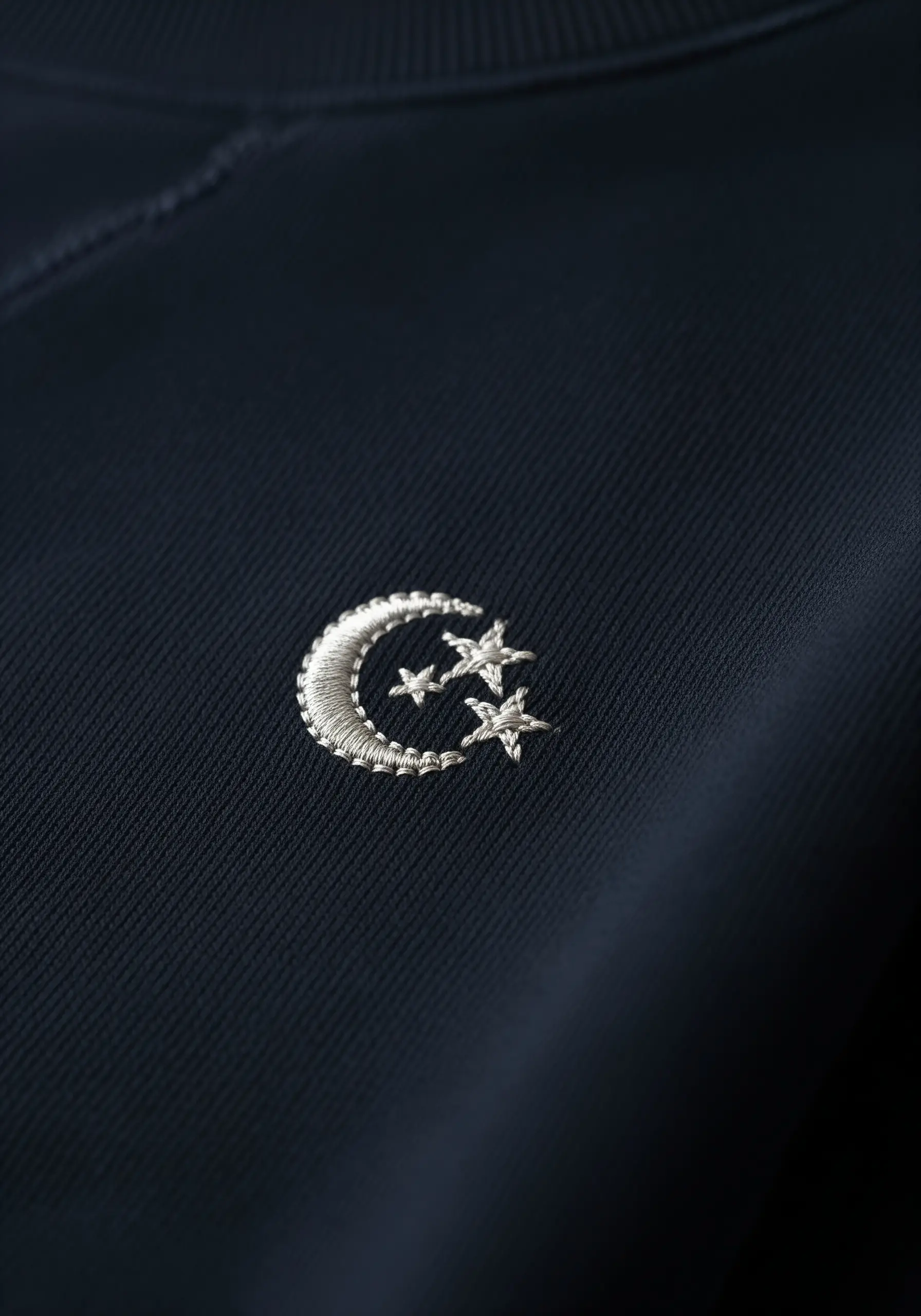A small, silver metallic crescent moon and stars embroidered on a navy blue sweatshirt.