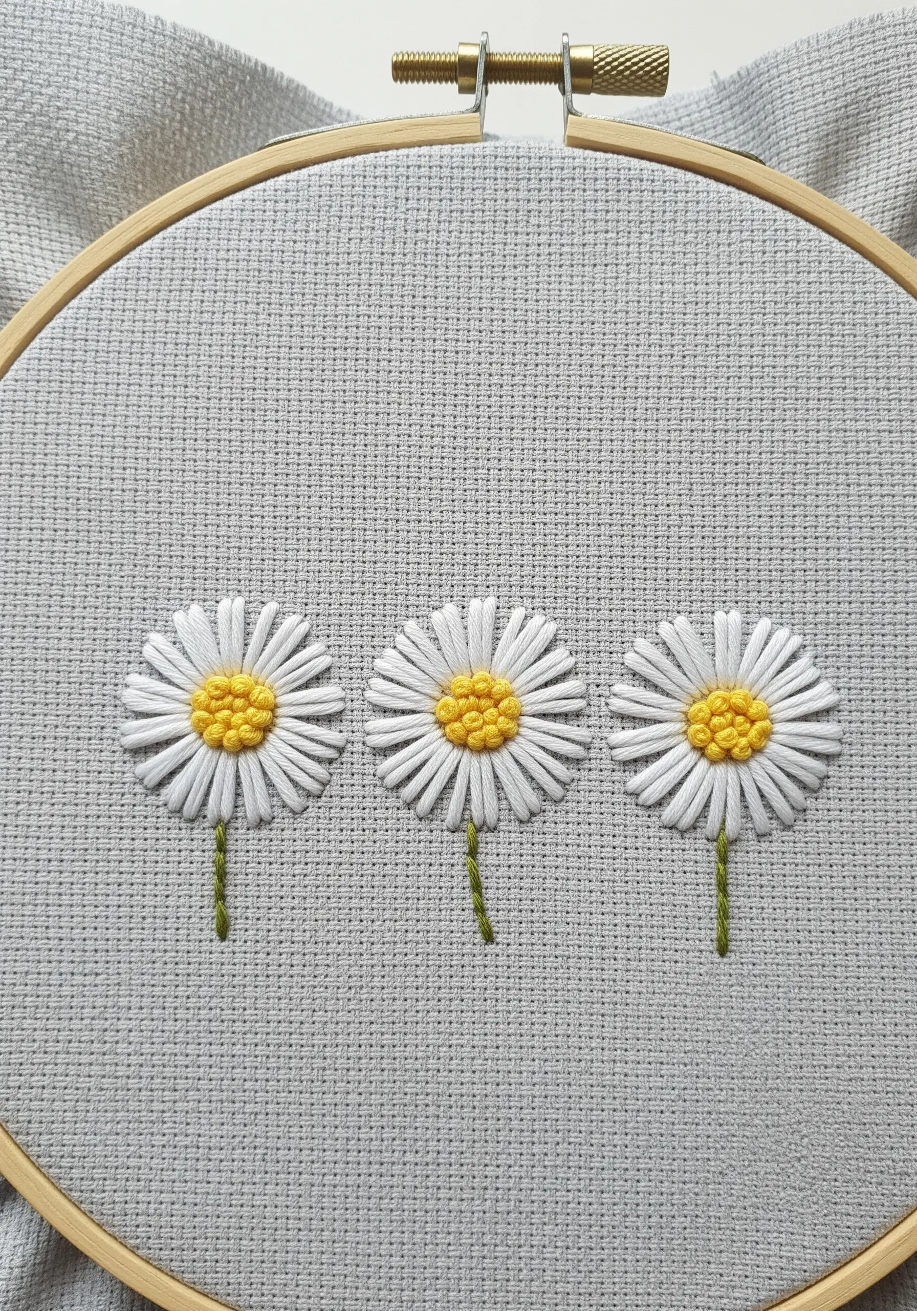 Three embroidered daisies in a row on light gray Aida cloth in a wooden hoop.