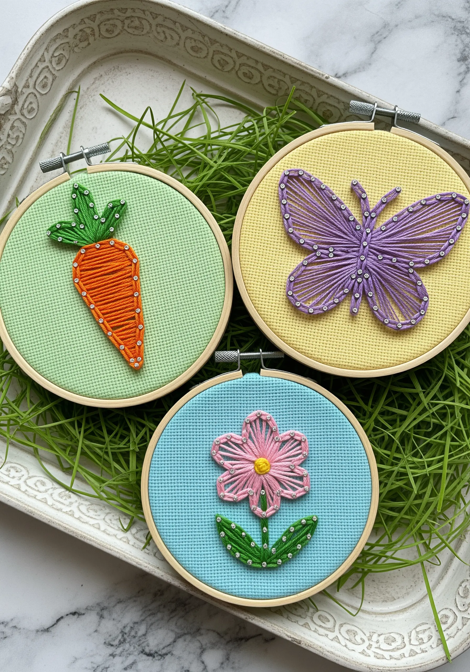 Three small string art hoops on gridded Aida cloth: carrot, butterfly, and flower.