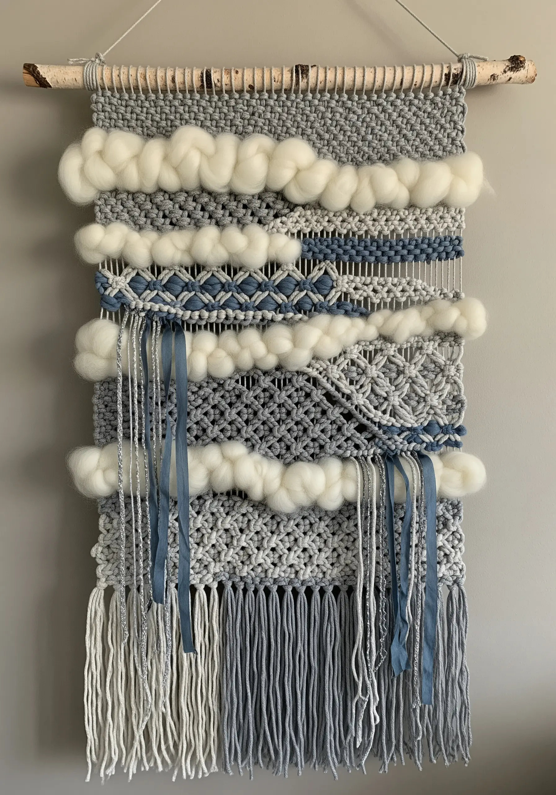 Woven wall hanging with chunky wool roving, macrame knots, and blue ribbon details on a birch branch.