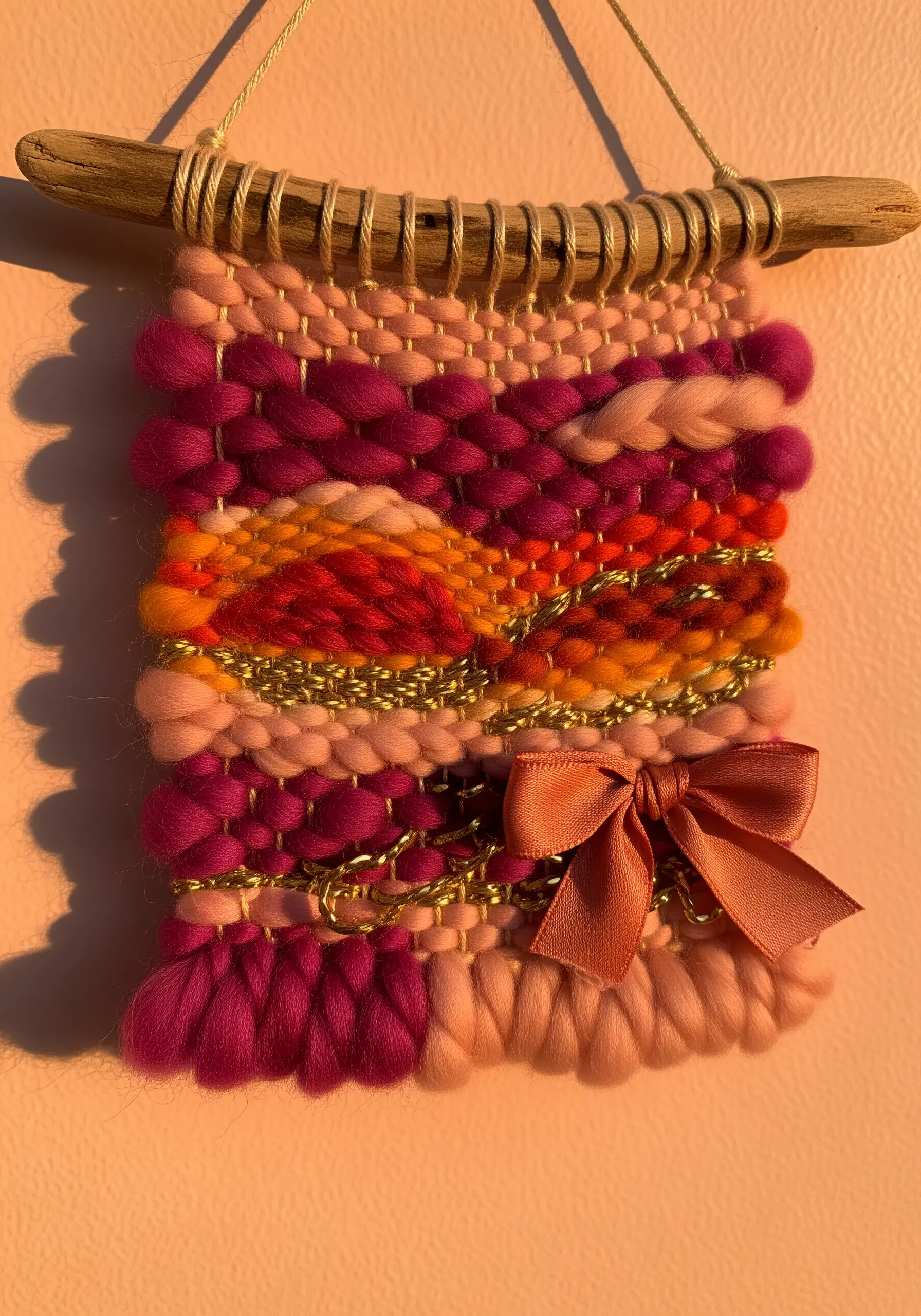 A small, colorful woven wall hanging with pink, orange, and gold tones on a driftwood branch.