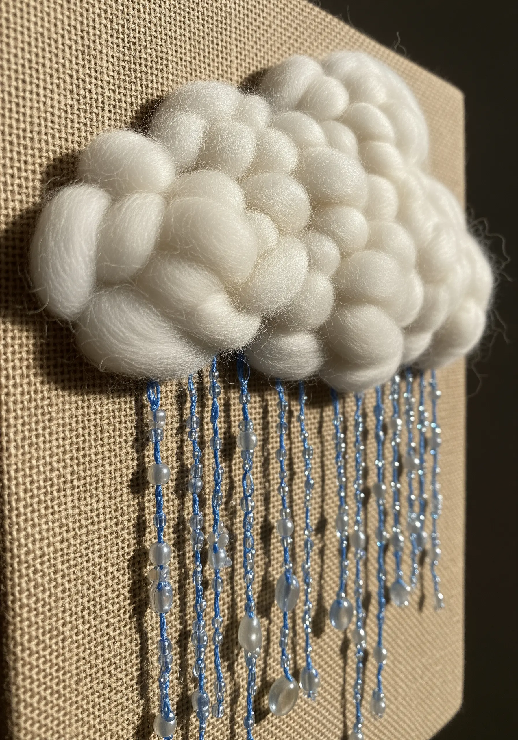 A textural cloud made from woven wool roving on burlap, with hanging beaded raindrops.