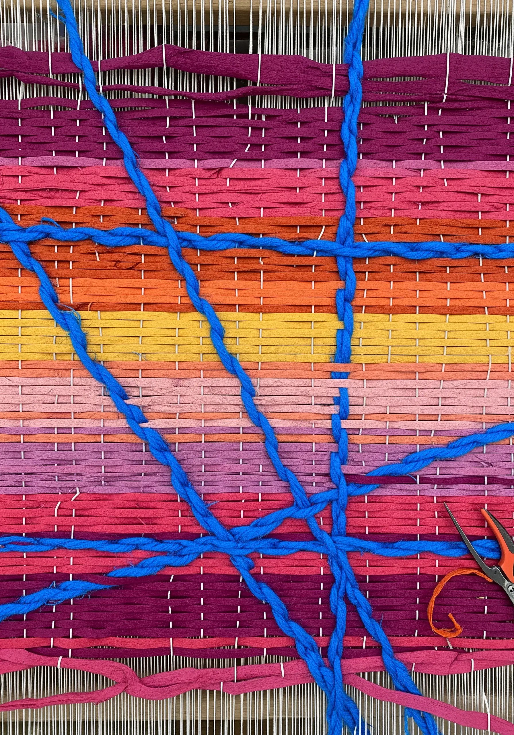 A close-up of a weaving made with colorful paper strips and thick blue braided yarn.